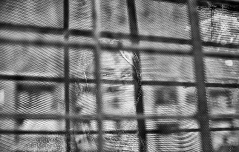 L'image montre une femme qui apparaît derrière une grille. Le contraste entre le flou et les barreaux de la grille donne une impression de séparation ou d'enfermement. La femme semble contemplative, et son expression peut évoquer des émotions profondes. Les nuances de noir et blanc ajoutent une atmosphère mélancolique à la scène.