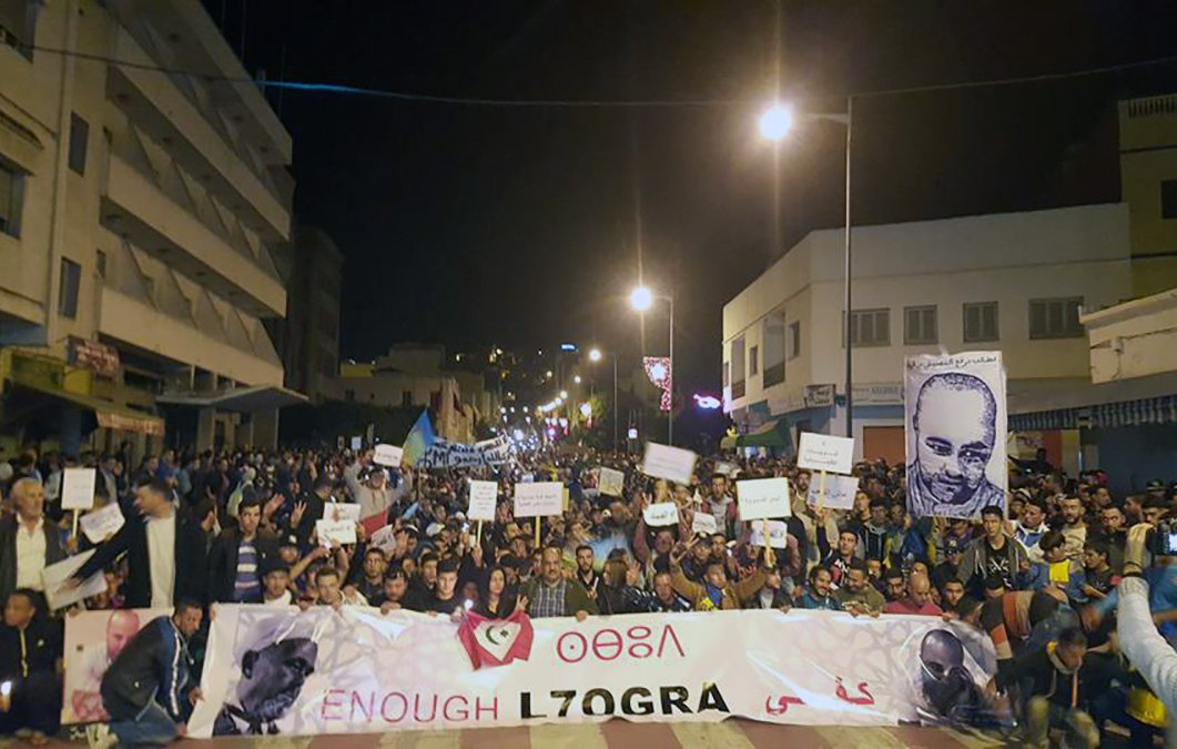 L'image montre une grande manifestation nocturne dans une rue animée. Beaucoup de personnes sont rassemblées, tenant des pancartes et banderoles. On peut voir des portraits de plusieurs individus exposés parmi les manifestants. L'atmosphère semble énergique et engagée, avec des participants qui expriment leurs revendications. Des lumières artificielles illuminent la scène, créant une ambiance dynamique.