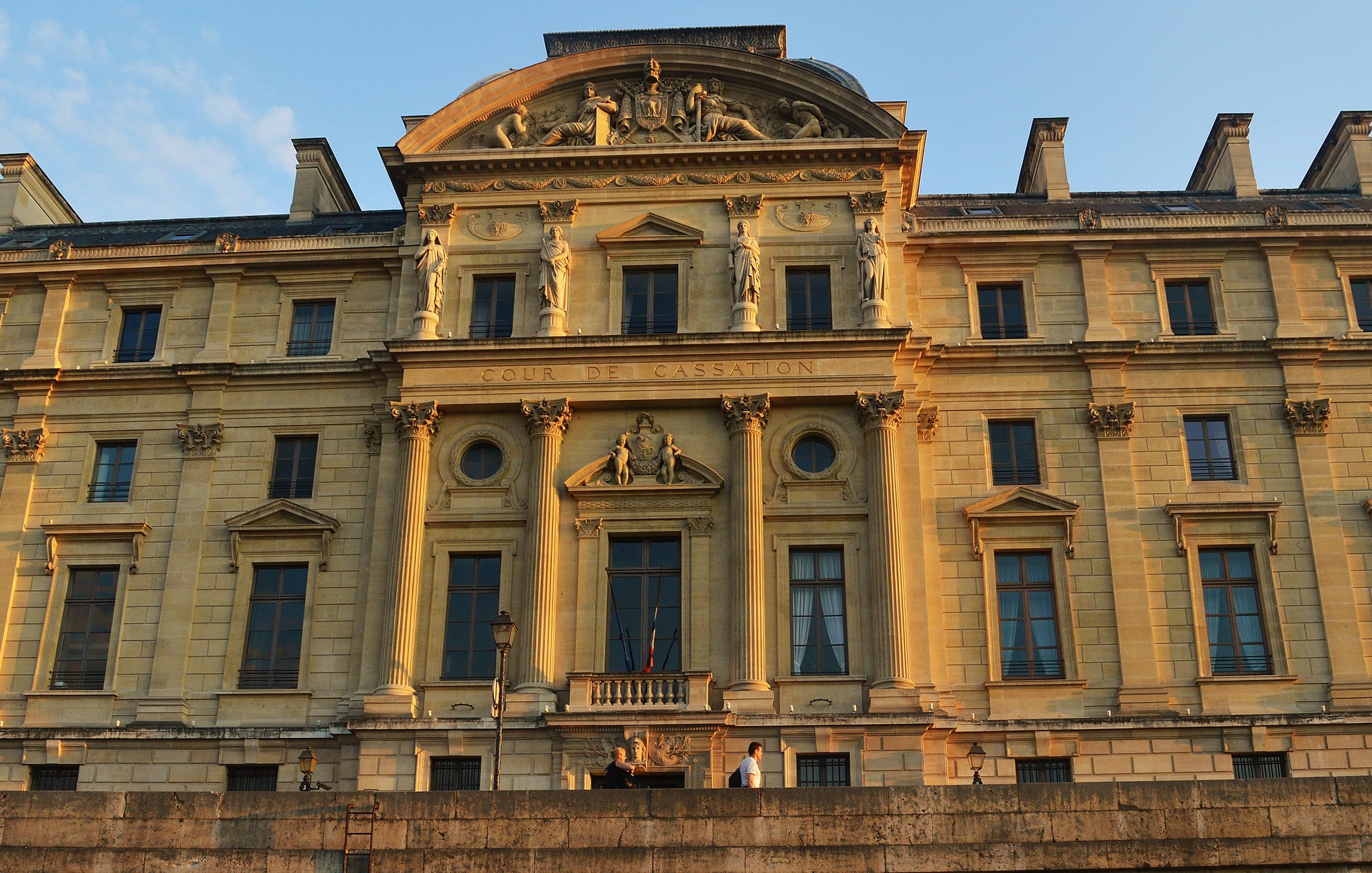 L'image montre un bâtiment imposant, probablement un palais ou un tribunal, éclairé par la lumière dorée du soleil couchant. La façade est ornée de détails architecturaux élaborés, avec des colonnes et des sculptures qui ajoutent à son majestueux style classique. Le nom "COUR DE CASSATION" est visible, indiquant qu'il s'agit d'un lieu judiciaire important. Le cadre montre un ciel clair, accentuant la beauté de l'architecture historique.