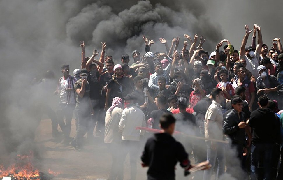 L'image montre un groupe de personnes rassemblées dans une atmosphère tendue. Elles sont entourées de fumée noire, probablement causée par des feux. Certains manifestants lèvent les bras en signe de protestation. On peut sentir une ambiance de colère et de résistance, avec quelques personnes qui semblent brandir des objets. Le décor est chaotique, évoquant une situation de crise ou de manifestation.