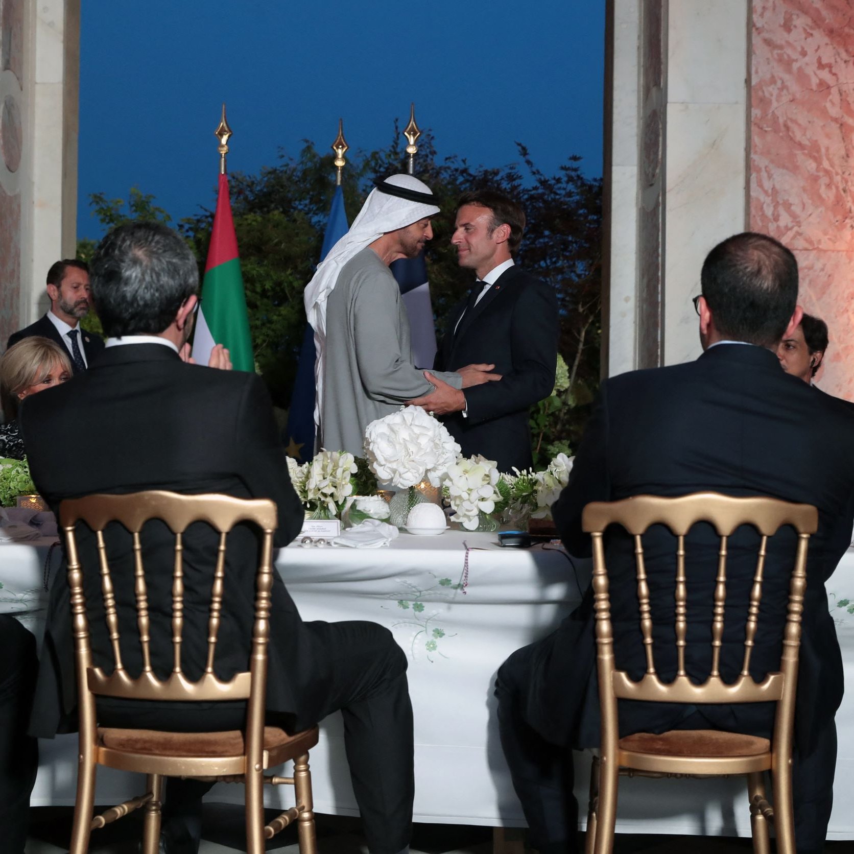 تظهر الصورة مجموعة من الأشخاص في مناسبة رسمية، حيث يقف رجلان في المقدمة يتبادلان التحية. أحدهم يرتدي غطاء رأس عربي، بينما الآخر يرتدي بدلة رسمية. في الخلفية، يوجد علم الإمارات العربية المتحدة وعلم آخر. يجلس الباقون حول طاولة مزينة بالزهور، حيث يبدو عليهم التفاعل مع الموقف. الإضاءة تعكس أجواء المساء، مما يضفي لمسة مميزة على الصورة.