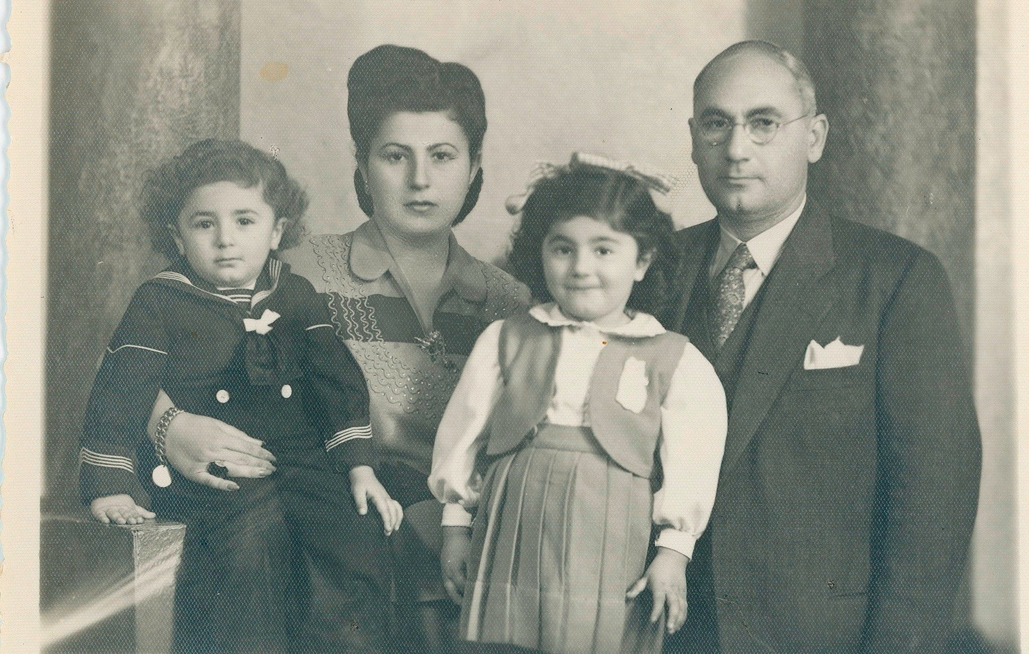Cette image en noir et blanc montre une famille posant ensemble. À gauche, un jeune garçon portant une tenue de marin est assis. À côté de lui, une femme au style classique avec des cheveux coiffés en pompadour se tient. En face d'elle, une petite fille avec un sourire doux porte une robe avec un gilet. À droite, un homme en costume, portant des lunettes, regarde la caméra. L'arrière-plan est sobre, ajoutant une touche nostalgique à cette photo de famille.
