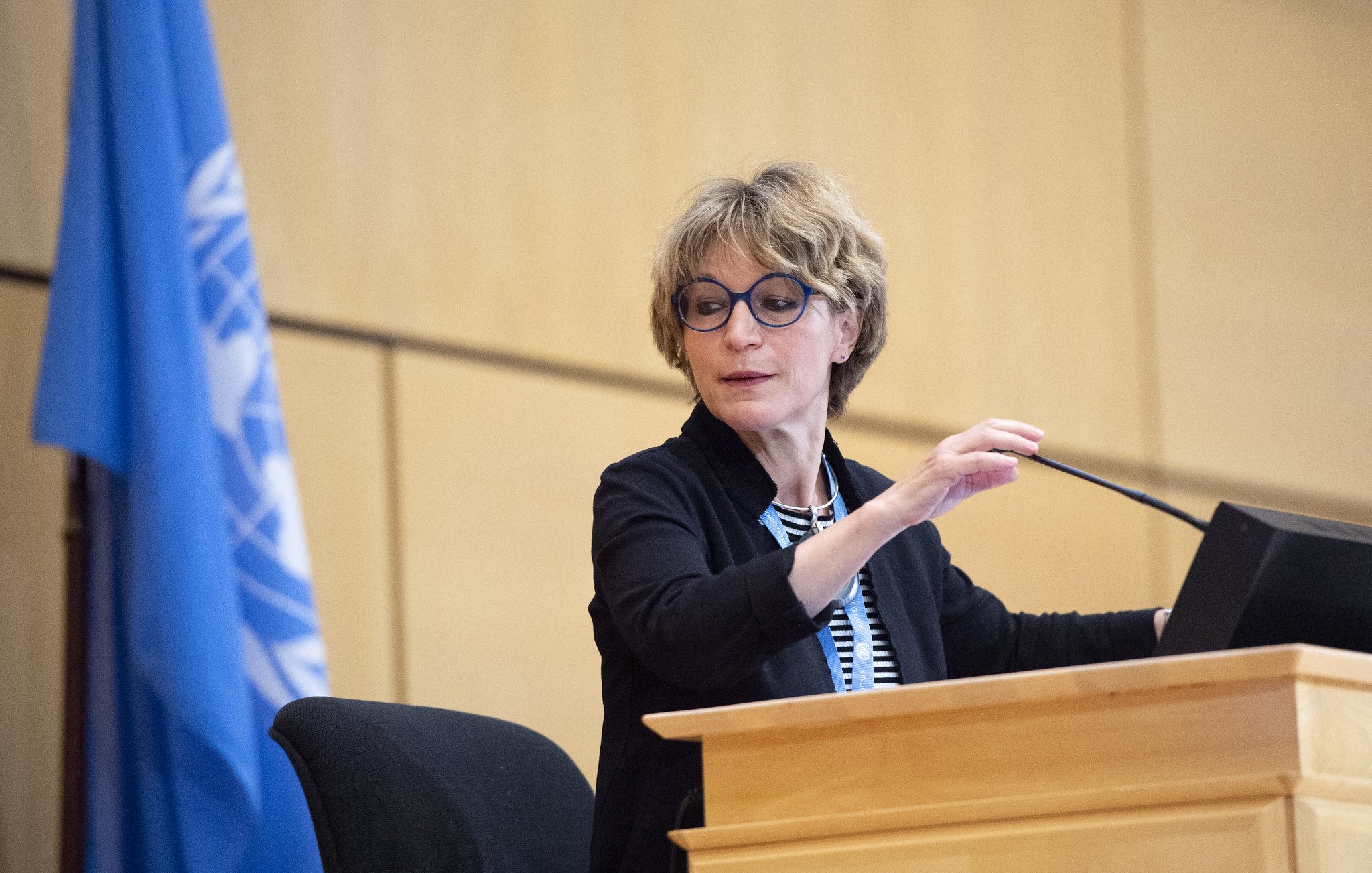 Femme avec des lunettes, parlant devant un podium, drapeau de l'ONU derrière elle.