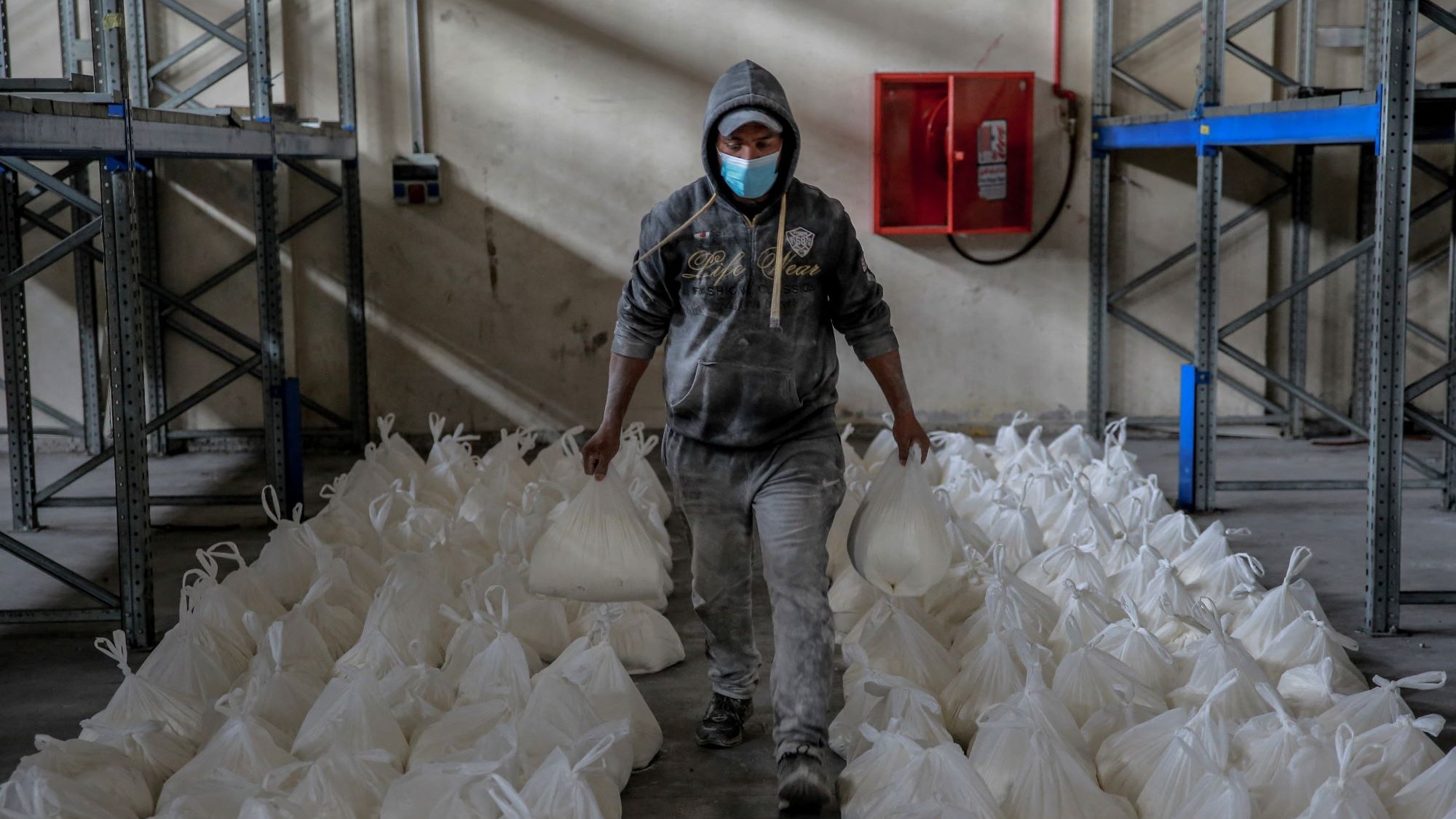 L'immagine mostra un uomo che cammina in un ambiente di magazzino. Indossa una felpa con cappuccio e una mascherina e tiene in mano due sacchetti bianchi. Intorno a lui ci sono numerosi sacchetti bianchi disposti ordinatamente sul pavimento. Le pareti del magazzino sono spoglie e ci sono scaffali metallici sullo sfondo. L'atmosfera sembra lavorativa e organizzata.