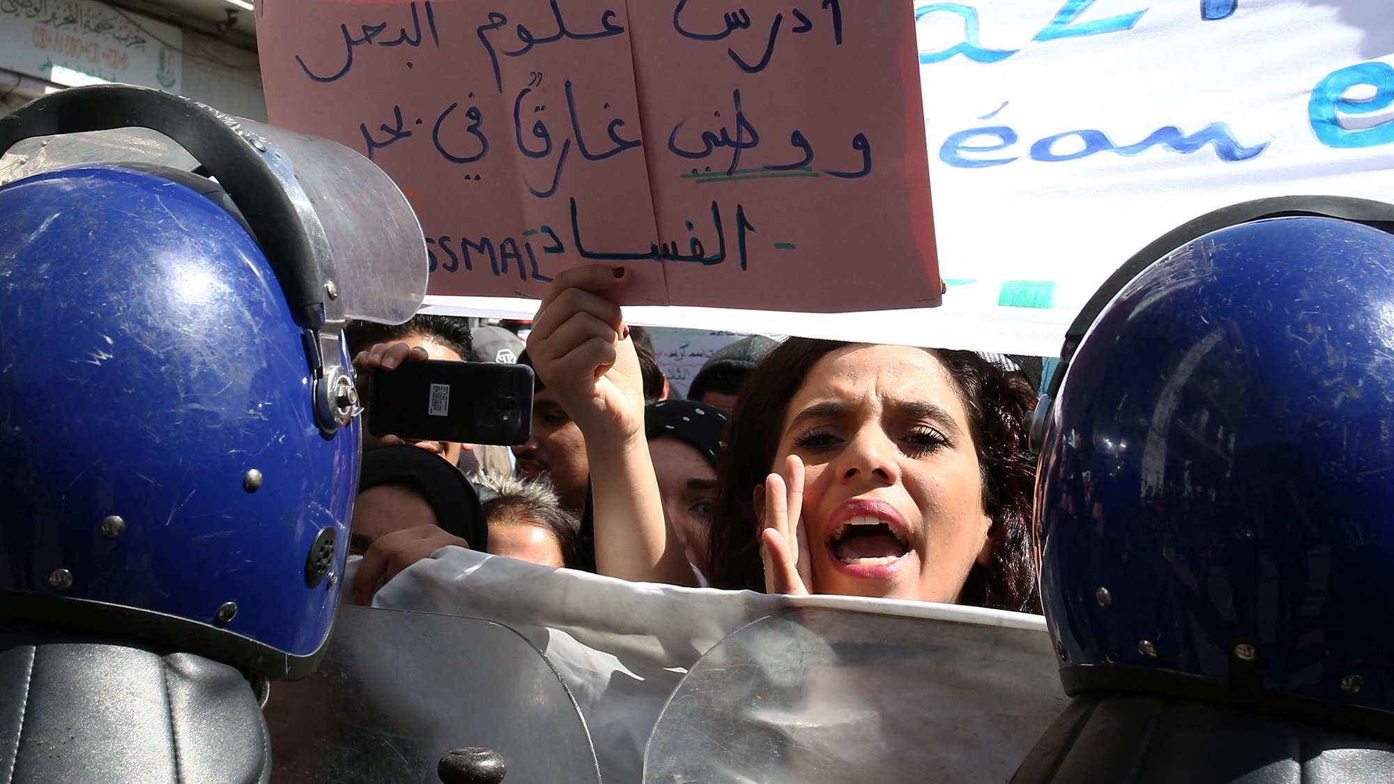 L'image montre une scène de manifestation où des personnes se rassemblent. À l'avant-plan, une femme semble exprimer des émotions vives, probablement en train de parler ou de crier, tandis qu'elle tient une pancarte. Plusieurs agents de la police, en uniforme avec des casques, se tiennent en ligne, formant un barrage. En arrière-plan, on peut apercevoir d'autres manifestants et une bannière portée par la foule. L'atmosphère semble tendue et engagée, avec une forte volonté de faire entendre des revendications.