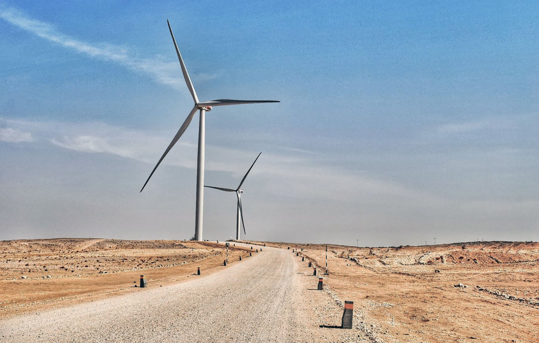 L'image montre un paysage désertique avec une route en gravier qui s'étend vers l'horizon. De chaque côté de la route, on aperçoit plusieurs éoliennes qui se dressent majestueusement contre un ciel bleu. Le terrain est aride, et les éoliennes symbolisent l'énergie renouvelable dans cet environnement désertique. Les colonnes le long de la route semblent délimiter le chemin, ajoutant une touche de structure à ce paysage naturel et technologique.