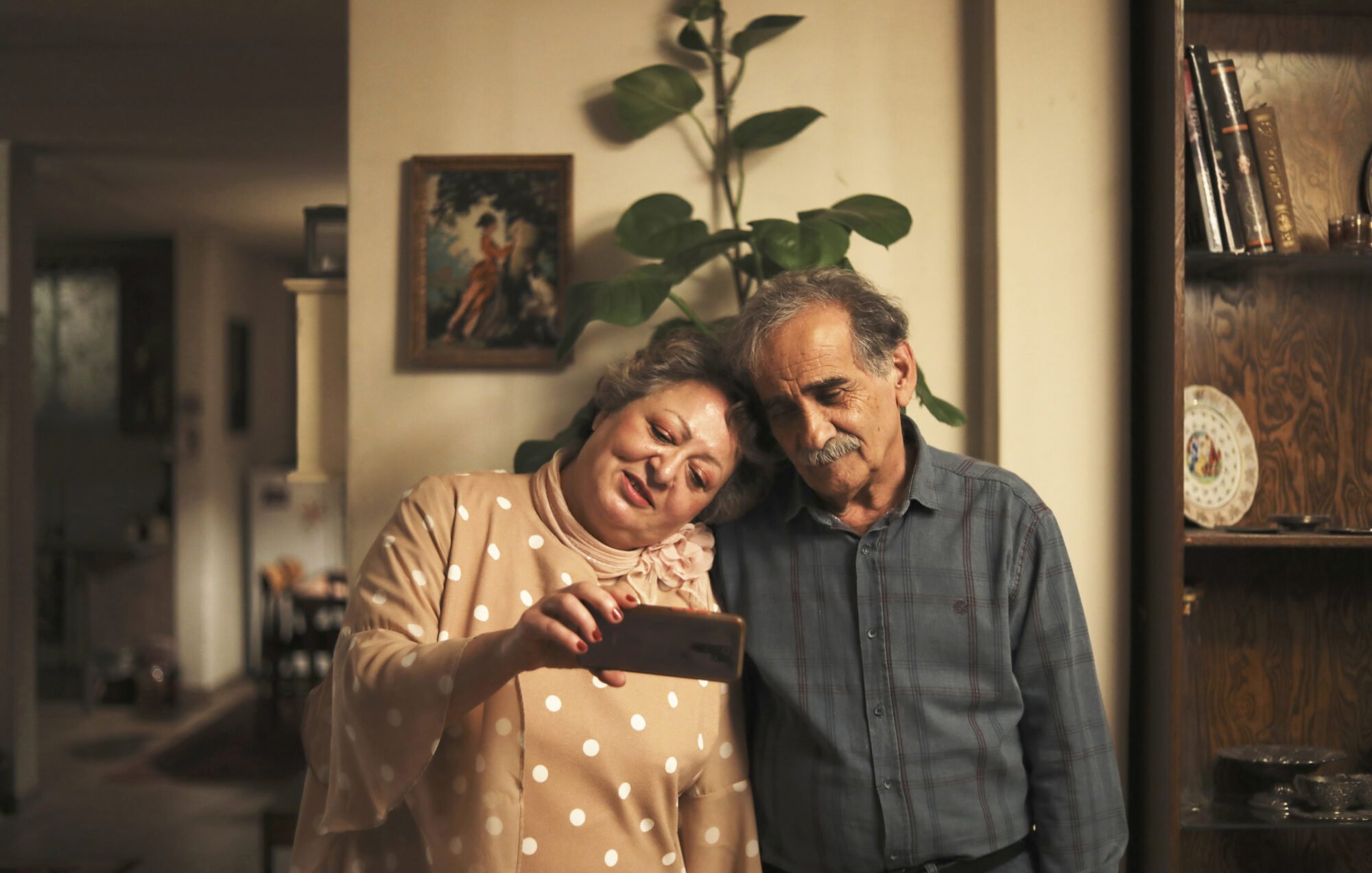 در این تصویر، یک زن و یک مرد در یک محیط داخلی ایستاده‌اند. زن لباسی با دکمه‌های بزرگ و کلاهی زیبا به تن دارد و در حال نشان دادن چیزی به مرد با گوشی همراه است. مرد در کنار او ایستاده و به صفحه گوشی نگاه می‌کند. هر دو لبخند بر لب دارند و به نظر می‌رسد که لحظه‌ای خوشحال‌کننده را تجربه می‌کنند. پس‌زمینه تصویر نشان‌دهنده یک خانه گرم و دلنشین است با گیاهانی در کنار دیوار.