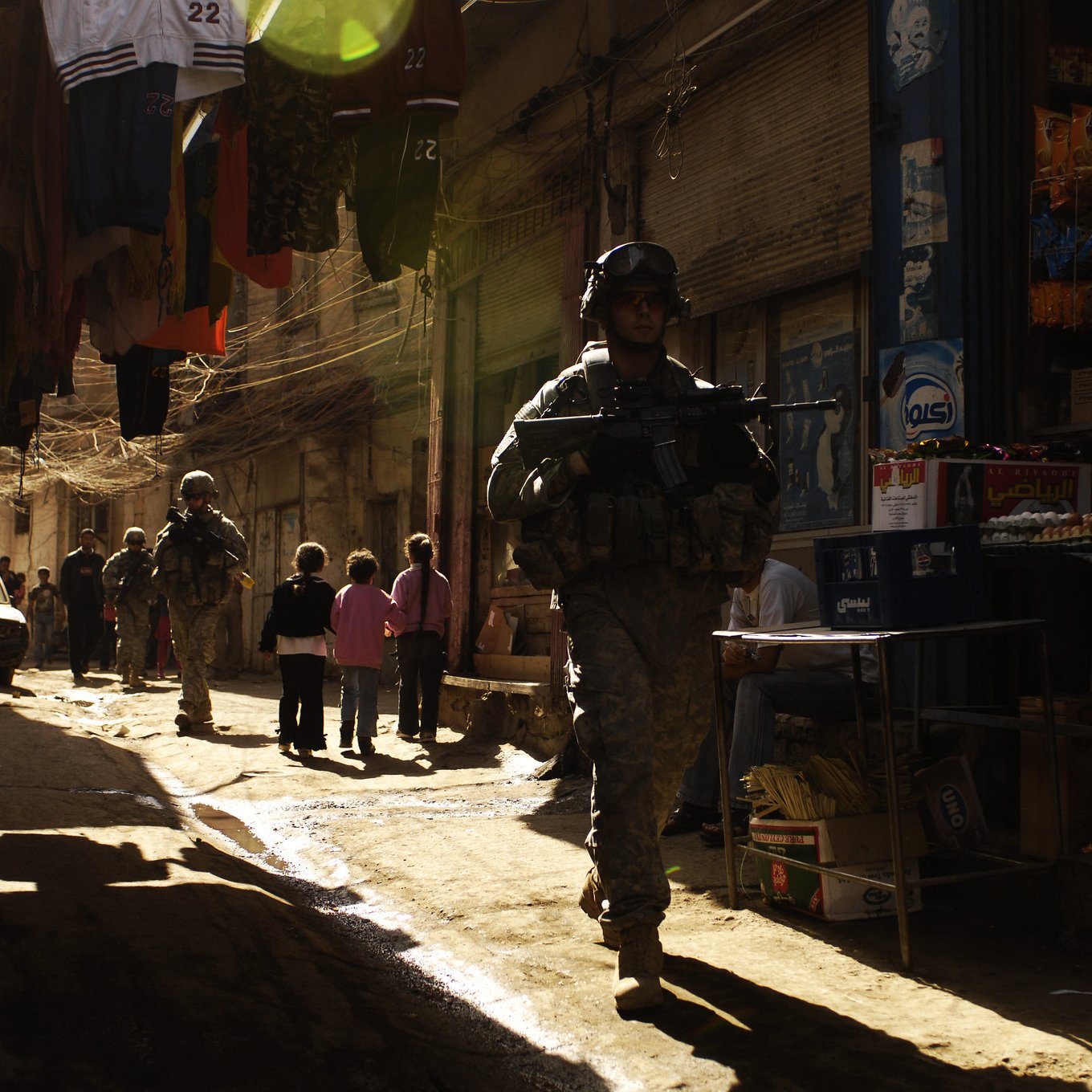 L'image montre une rue étroite et animée typique d'un marché urbain. Des soldats en uniforme, équipés d'armes, marchent le long de la ruelle. On aperçoit également des enfants qui semblent jouer ou observer les soldats. Les bâtiments sont proches les uns des autres, et des vêtements sont suspendus au-dessus de la rue. La lumière du soleil filtre à travers la ruelle, créant une ambiance contrastée entre l'ombre et la lumière. L'environnement semble à la fois vivant et chargé de tension.