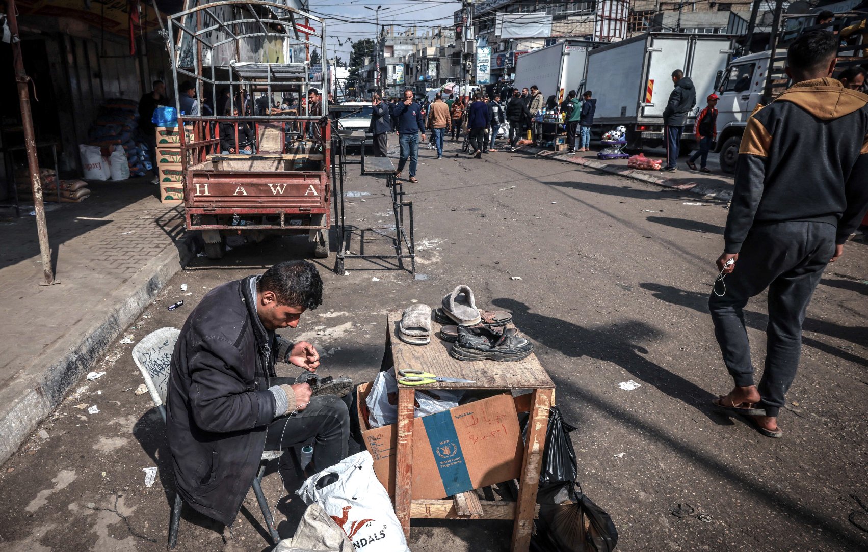 Nell'immagine c'è una strada affollata con diverse persone che si muovono. In primo piano, un uomo è seduto su una sedia accanto a un tavolo, occupato a riparare delle scarpe. Attorno a lui ci sono alcuni oggetti e sacchetti. Sullo sfondo si vedono veicoli e altre persone che camminano. L'atmosfera è vivace e caotica.