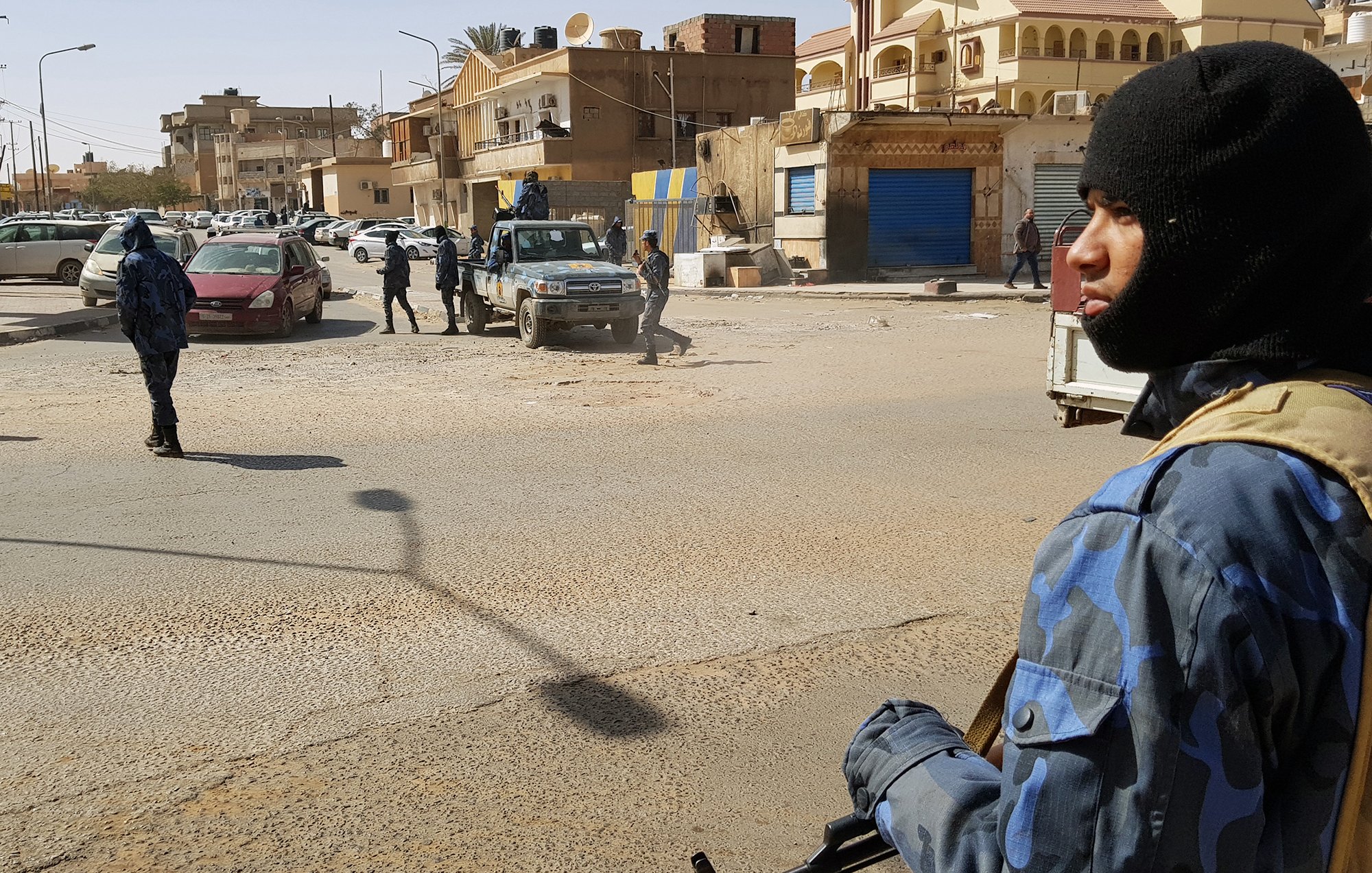 L'immagine mostra una scena di una zona urbana, presumibilmente in un contesto di sicurezza elevata. In primo piano, un uomo indossa un berretto e abbigliamento camuffato, mentre tiene un'arma. Sullo sfondo si possono vedere altri uomini in uniformi simili che si muovono in un'area in cui ci sono veicoli parcheggiati. L'architettura circostante appare tipica di una città, con edifici vari e attività commerciali. La luce del giorno illumina la scena, creando ombre ben definite.