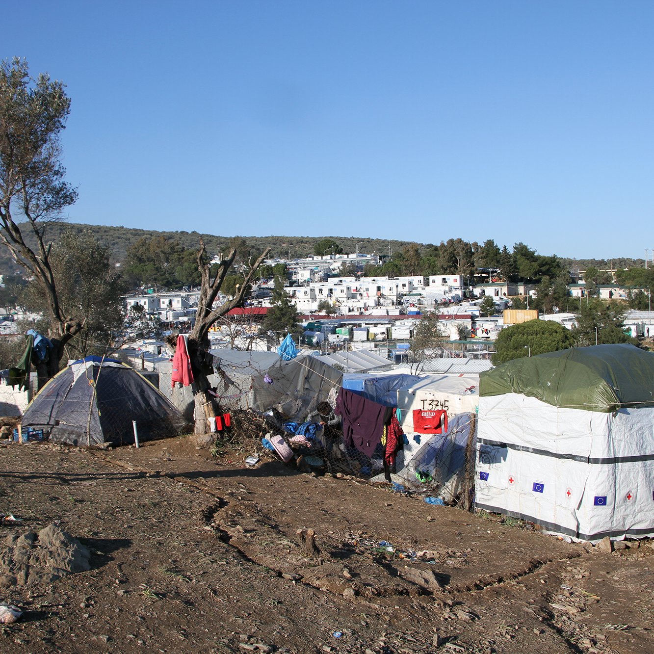L'image montre une scène d'un campement de migrants. On peut voir des tentes montées sur un terrain plutôt nu et chaotique, avec des déchets éparpillés autour. À l'arrière-plan, des bâtiments sont visibles, suggérant qu'il y a une communauté près de ce camp. Le ciel est clair et ensoleillé, et il y a quelques arbres et collines dans le paysage. Une personne se tient debout dans la scène, semblant observer les environs. L'ensemble dégage une impression de précarité et de lutte.