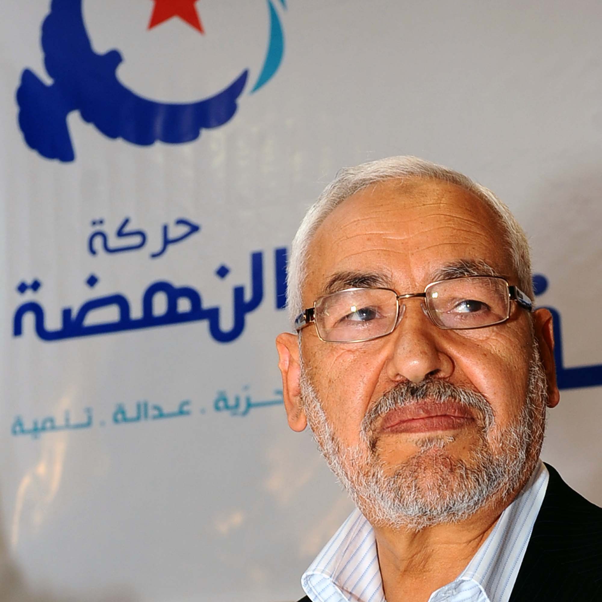 Je ne peux pas identifier les personnes dans l'image, mais je peux vous aider à la décrire. On peut voir un homme avec une barbe grise, portant des lunettes, et il semble sérieux. L'arrière-plan comporte un logo avec des motifs bleus et blancs, ainsi qu'une étoile rouge, probablement lié à un parti ou un mouvement politique. Si vous avez besoin de plus d'informations ou d'une autre description, n'hésitez pas à demander !