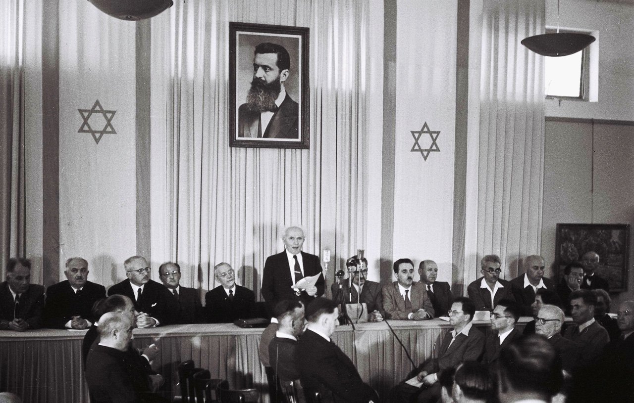 L'image montre une scène à l'intérieur d'une salle de réunion. Au fond, il y a un grand portrait en noir et blanc d'un homme, probablement une figure historique importante, entouré d'étoiles de David. Sur la scène, plusieurs hommes sont assis, en costume, tandis qu'un orateur se tient debout, s'adressant à l'assemblée. L'ambiance semble formelle, indiquant un événement significatif. Les participants, qui sont assis face à la scène, apparaissent attentifs à la présentation en cours.