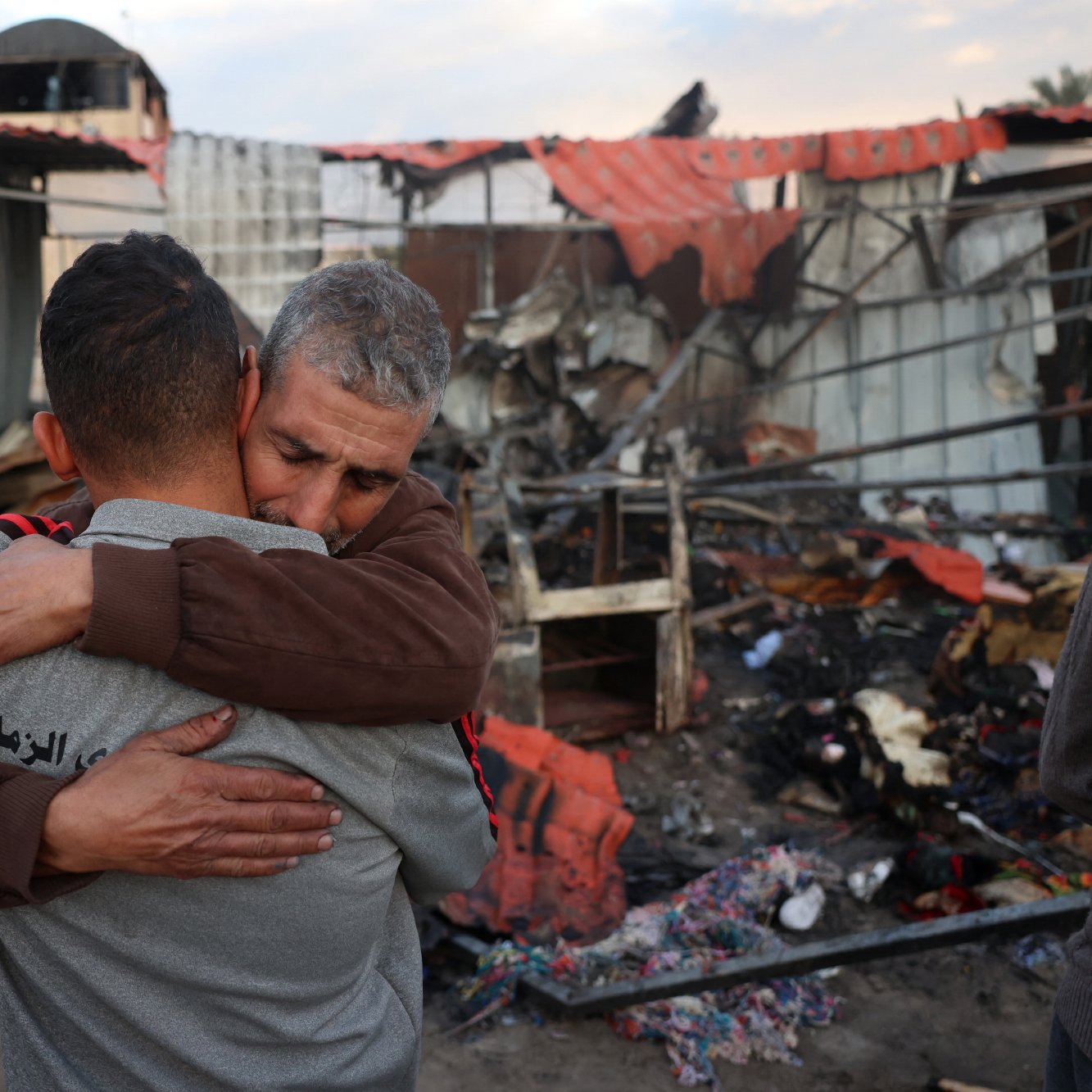 در این تصویر، دو مرد به یکدیگر در آغوش می‌افتند. پس‌زمینه تصویر نشان‌دهنده یک محل ویران شده است که به نظر می‌رسد دچار آتش‌سوزی یا تخریب شده است. حس ناراحتی و همدردی در چهره‌های آنها مشهود است و فضای کلی تصویر احساساتی و غم‌انگیز به نظر می‌رسد.