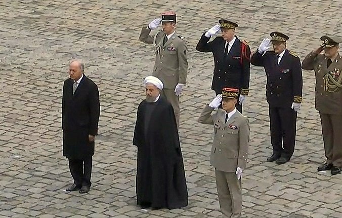 L'image montre plusieurs personnes en uniforme militaire saluant. Au centre, deux hommes en costume : l'un à gauche semble être un dirigeant, tandis que l'autre, portant une robe noire et un turban, est probablement un haut responsable religieux ou politique. Les autres personnes en arrière-plan sont des officiers en uniforme, également en train de rendre les honneurs. Ils se trouvent sur une surface en pavés, suggérant une cérémonie officielle ou un événement diplomatique.