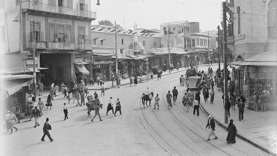 L'image montre une vue animée d'une rue commerçante dans une ville. On y voit des bâtiments à plusieurs étages avec des balcons, des étals et des commerces le long de la rue. De nombreuses personnes, y compris des piétons et des animaux, circulent. On aperçoit également une voiture ancienne sur la route. L'atmosphère semble vibrante et typique d'une époque passée, avec un mélange de textures urbaines et de la vie quotidienne.
