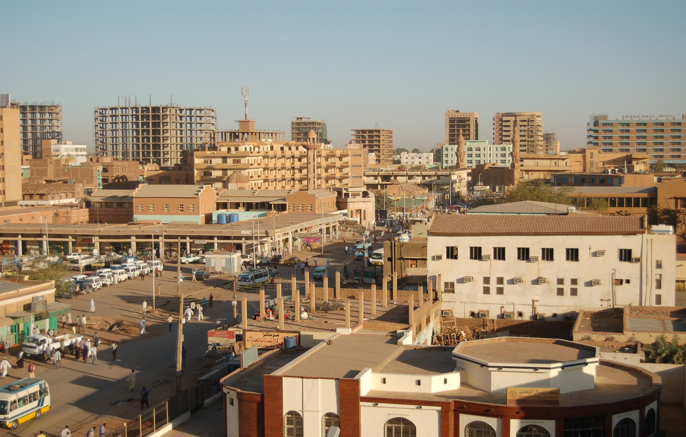 الصورة تظهِر مشهداً حضرياً لمدينة تحتوي على مبانٍ متعددة الطوابق، وبعض المباني قيد الإنشاء. تُظهر الشوارع المزدحمة بالسيارات والمارة، بالإضافة إلى وجود أسواق محلية وأعمال تجارية. السماء صافية، وهناك ضوء شمس ساطع، مما يوحي بأن الوقت نهار. الألوان في الصورة تتراوح بين درجات البني والرمادي، مما يعكس طابع المدينة.