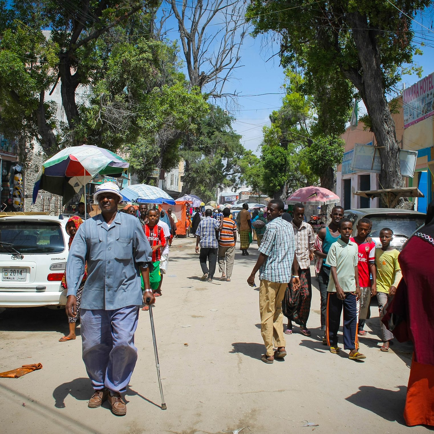 L'immagine mostra una strada affollata, probabilmente in un'area urbana. Ci sono diversi gruppi di persone che camminano, alcune sotto ombrelloni. Un uomo anziano con una canna si muove lentamente, mentre altri sembrano conversare e fare acquisti. Sullo sfondo, si possono vedere alcune automobili parcheggiate e alberi che offrono ombra. L'atmosfera sembra vivace e comunitaria, con il sole che illumina la scena.