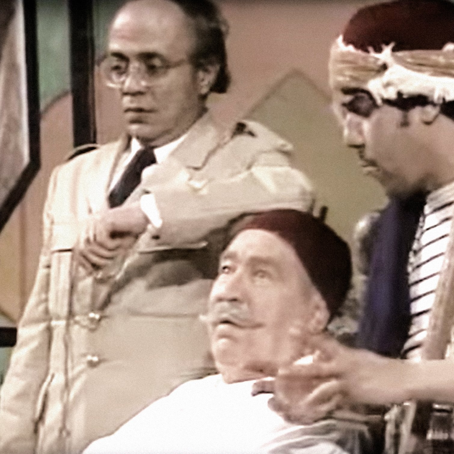 L'image montre trois personnages dans un cadre qui semble être une scène de théâtre ou une comédie. L'un des personnages est assis, portant une blouse blanche et un fez, affichant une expression détendue. À sa gauche se trouve un personnage en tenue militaire, regardant sa montre avec une expression préoccupée. À droite, un troisième personnage, également en fez, joue d'un instrument de musique tout en gesticulant, ajoutant une ambiance dynamique à la scène. L'ensemble évoque un moment comique ou dramatique.