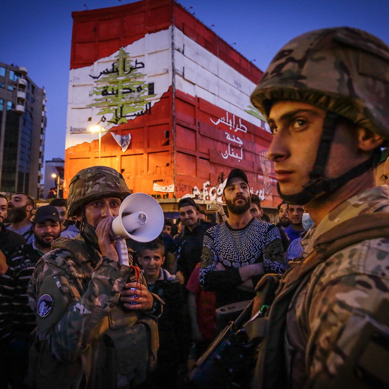 The image depicts a scene of a gathering in an urban environment during the evening. In the foreground, soldiers in military gear, including helmets, are present, with one holding a megaphone, suggesting a communication or announcement to the crowd. The background features a large, colorful wall or mural that displays the Lebanese flag and some Arabic text, likely conveying a political or social message. The crowd surrounding the soldiers appears engaged, with various expressions on their faces, indicating a sense of tension or anticipation in the atmosphere. The setting is illuminated by artificial lighting, hinting at the late hour.
