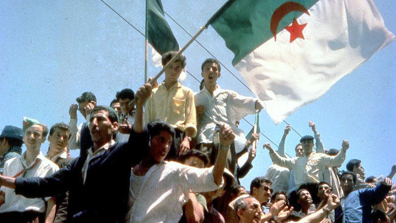 L'image montre une foule rassemblée, levant des bras et brandissant des drapeaux, dont celui de l'Algérie. Les personnes semblent joyeuses et engagées, célébrant un moment fort. L'atmosphère pourrait évoquer une célébration de l'indépendance ou un événement historique significatif pour le pays. Les visages sont animés et l'énergie collective est palpable.