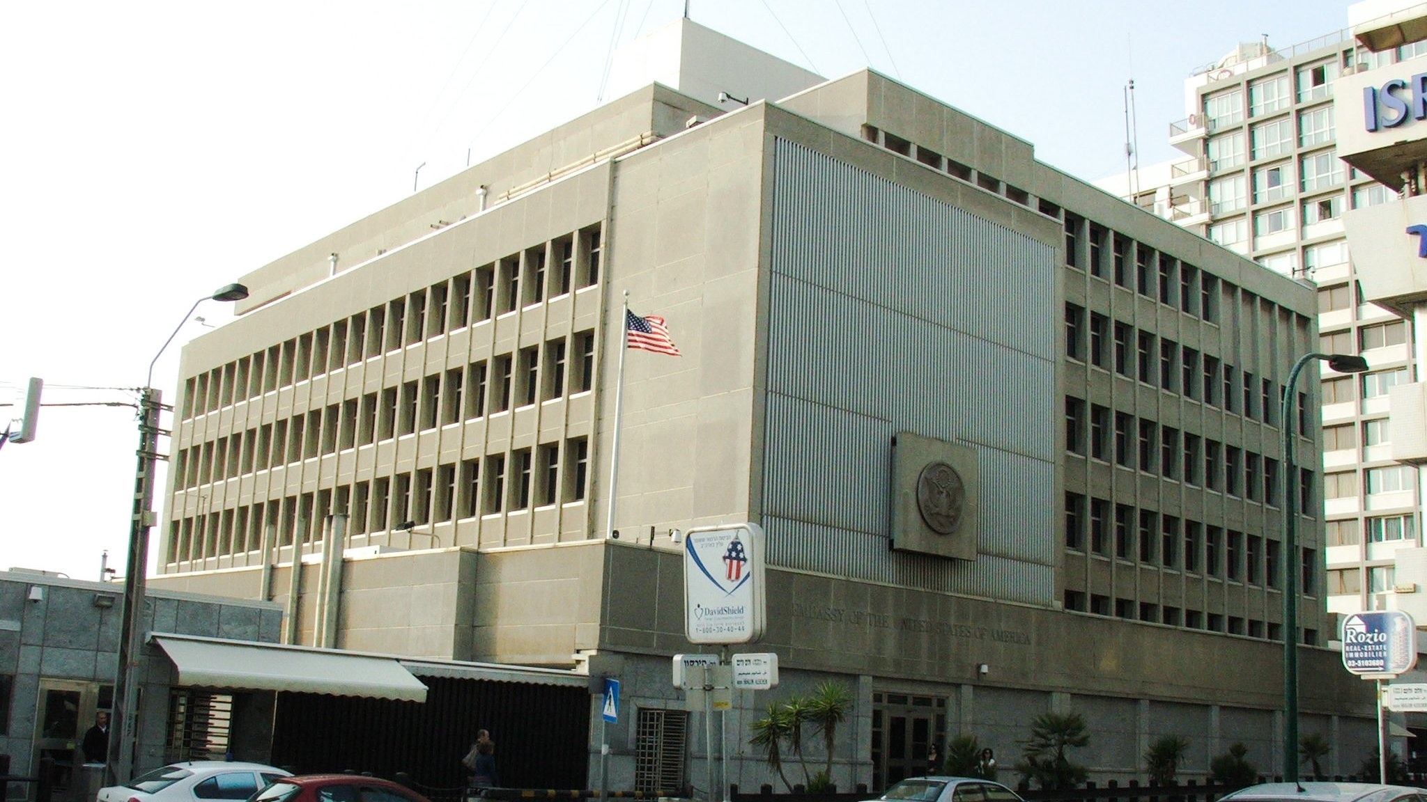 L'image montre un bâtiment moderne avec une architecture fonctionnelle. Il est en béton, avec plusieurs fenêtres rectangulaires sur la façade. Un drapeau américain est visible sur le toit, indiquant que c'est une représentation diplomatique. À l'avant du bâtiment, il y a des panneaux de signalisation et quelques véhicules garés le long de la route. L'environnement semble urbain, avec d'autres immeubles en arrière-plan.