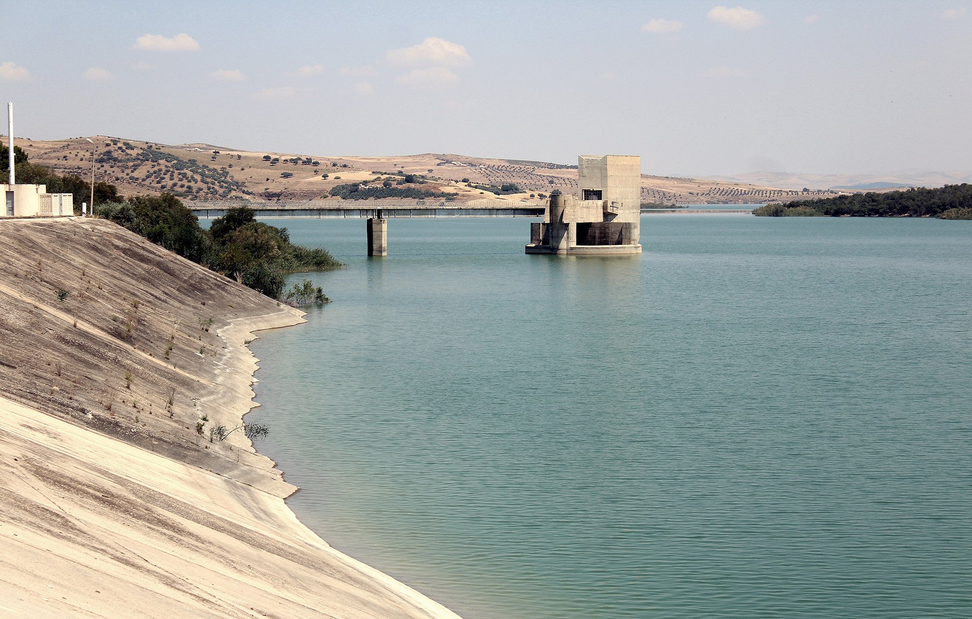 L'image montre un paysage calme avec un grand plan d'eau, probablement un réservoir ou un lac. Sur la gauche, la rive est recouverte d'une surface lisse et légèrement inclinée, tandis qu'à droite, on peut voir une structure en béton isolée au milieu de l'eau, avec des piliers qui émergent. À l'arrière-plan, des collines arrondies se dressent, ajoutant une touche de verdure au paysage. Le ciel est partiellement nuageux, ce qui crée une atmosphère paisible et naturelle.