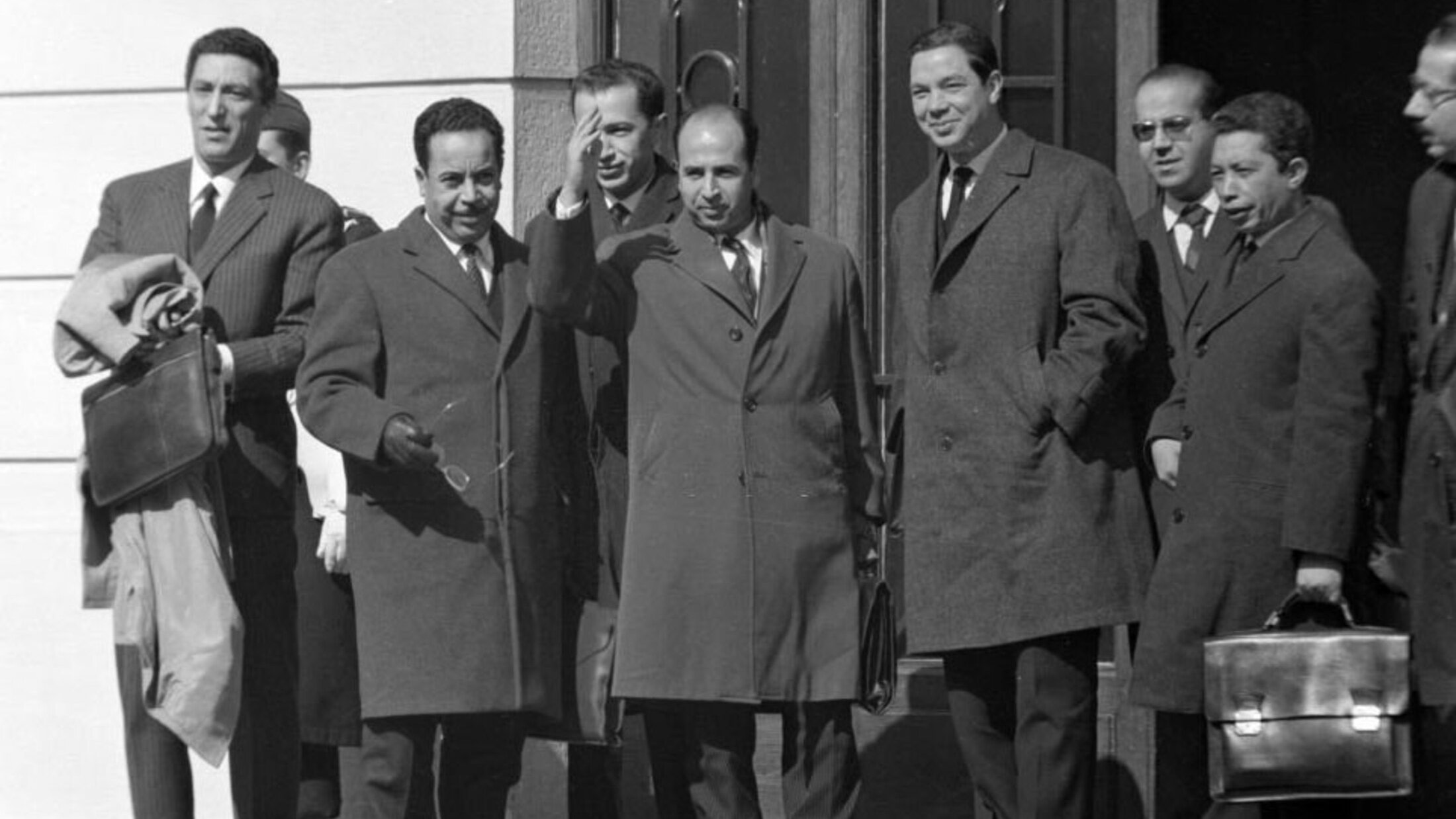 L'image montre un groupe de plusieurs hommes en costume, se tenant à l'extérieur d'un bâtiment. Ils portent des manteaux d'hiver, indiquant que la scène se déroule probablement par temps frais. L'un des hommes, au centre, semble être en train de saluer ou de parler avec enthousiasme. Les autres hommes affichent des expressions neutres ou souriantes. L'environnement est urbain, avec des détails architecturaux en arrière-plan.