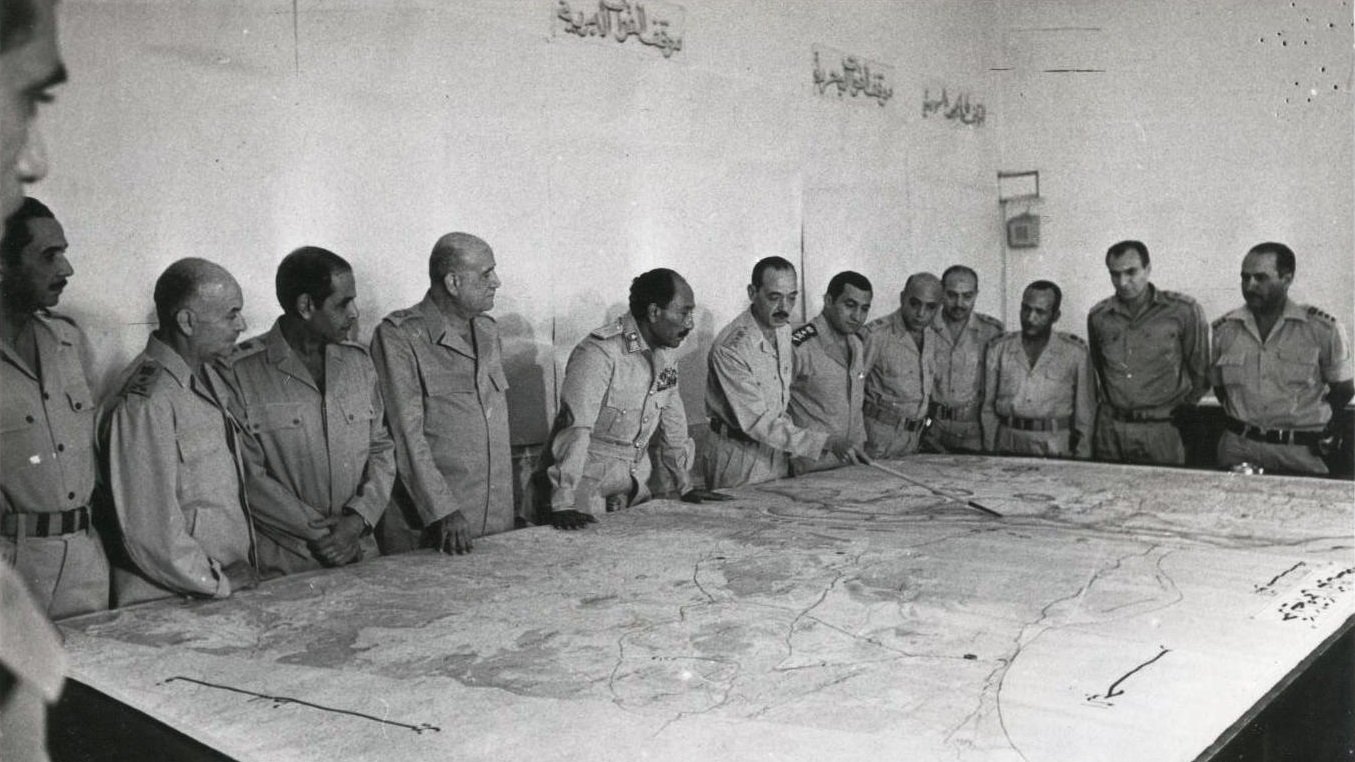 تظهر الصورة مجموعة من الرجال يرتدون زيًا عسكريًا، يجتمعون حول طاولة كبيرة تعرض خريطة. يبدو أن الاجتماع يدور حول التخطيط أو مناقشة استراتيجية ما. تعكس تعابير وجوههم التركيز والاهتمام بما يُناقش. الجدران خلفهم تبدو بسيطة، مع بعض الكتابات باللغة العربية. تُعطي الصورة انطباعًا عن موقف رسمي وعسكري.