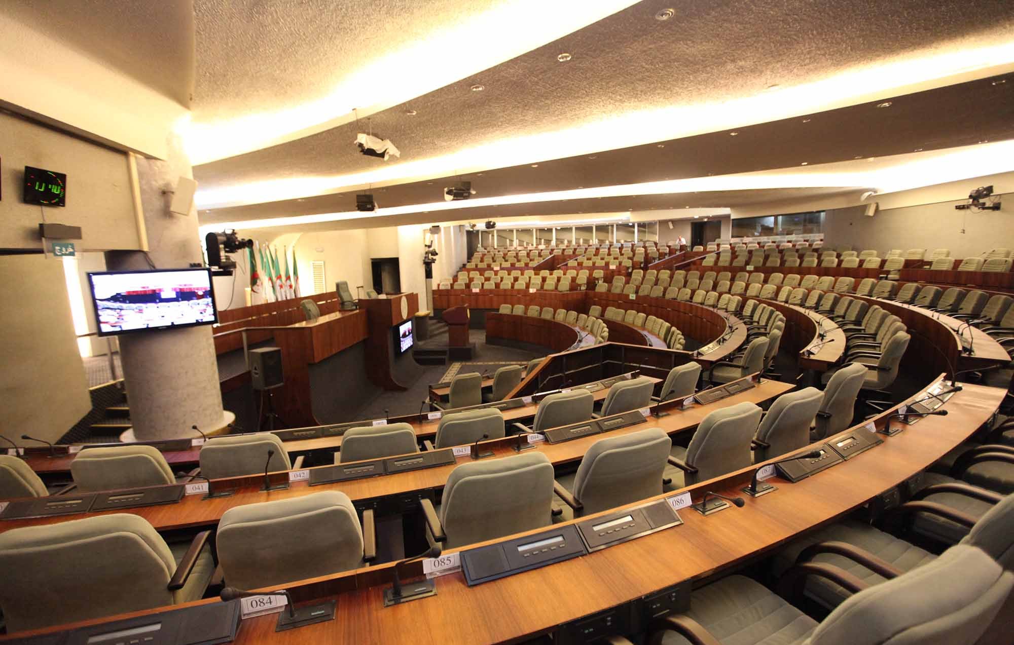 L'image représente une salle de réunion ou un hémicycle, typique des assemblées législatives. On peut voir des sièges disposés en amphithéâtre, avec des rangées de fauteuils gris. À l'avant, il y a une estrade avec un bureau et des équipements audiovisuels. La décoration est moderne et sobre, montrant un environnement formel adapté aux discussions et aux débats. Des écrans sont également présents pour fournir des informations aux membres présents.