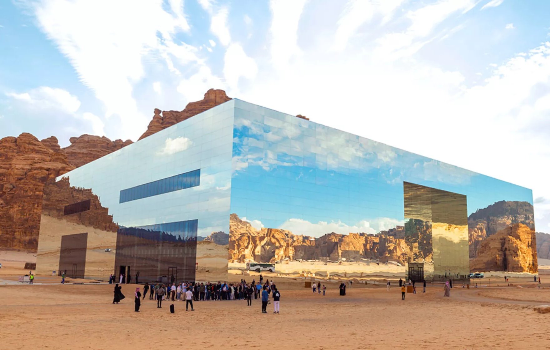 L'image montre un bâtiment moderne et futuriste situé dans un paysage désertique. La structure est entièrement recouverte de miroirs, reflétant le ciel et les montagnes environnantes, ce qui lui permet de se fondre harmoniellement dans le paysage. Au premier plan, un groupe de personnes, vraisemblablement des visiteurs, se regroupe sur le sable. L'atmosphère est paisible, et le ciel est parsemé de nuages, créant un contraste avec les teintes désertiques. L'architecture innovante et les réflexions ajoutent une dimension visuelle fascinante à l'environnement naturel.