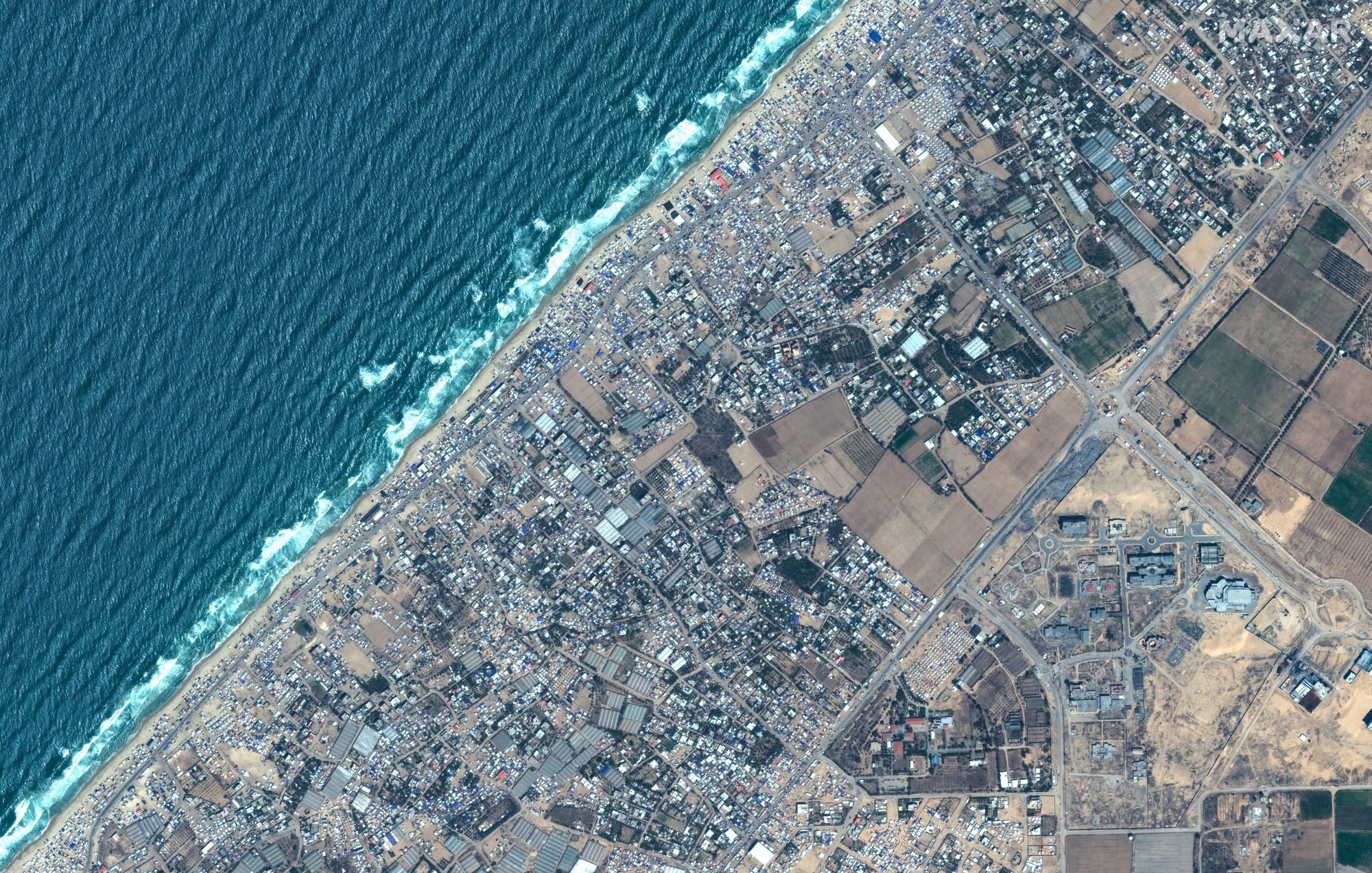 این تصویر یک نمای هوایی از ناحیه‌ای است که شامل سواحل و مناطق مسکونی و کشاورزی می‌باشد. در سمت چپ تصویر، یک نوار آبی رنگ نمایان است که به دریا اشاره دارد، در حالی که در سمت راست، زمین‌های کشاورزی و ساختمان‌های مسکونی وجود دارند. بافت شهری به صورت متراکم و پراکنده دیده می‌شود و همچنین برخی مناطق سبز و زمین‌های زراعی نیز در پایین تصویر مشخص است. 