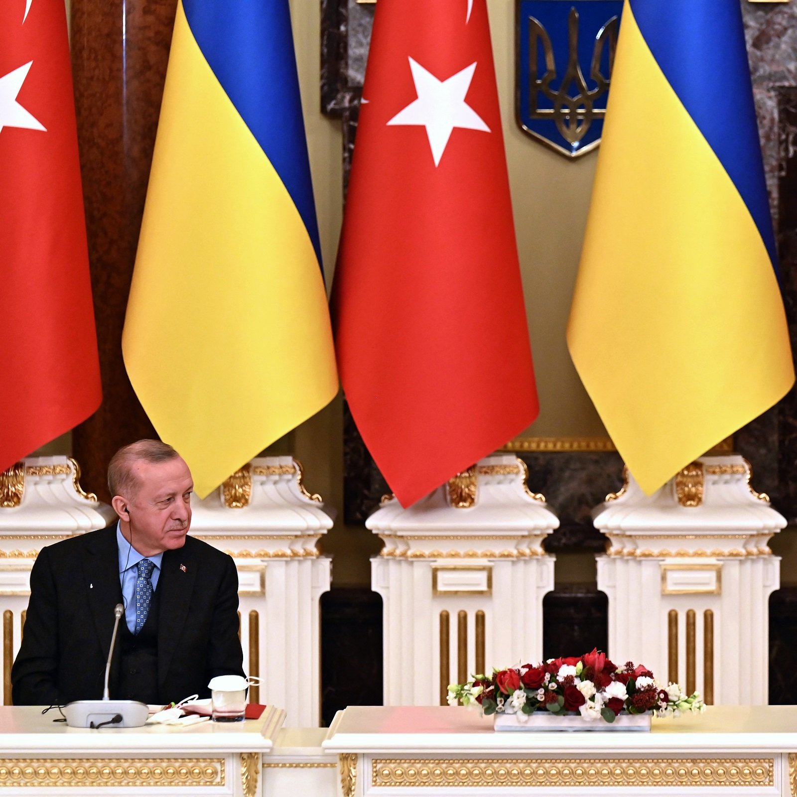 L'image montre une scène officielle avec deux hommes assis à une table. À gauche, un homme porte un costume sombre et semble en discussion. À droite, un autre homme est également en costume, regardant vers son interlocuteur. Derrière eux, on voit des drapeaux turcs et ukrainiens disposés de manière symétrique, et une décoration élégante. L'atmosphère générale évoque une rencontre diplomatique importante.