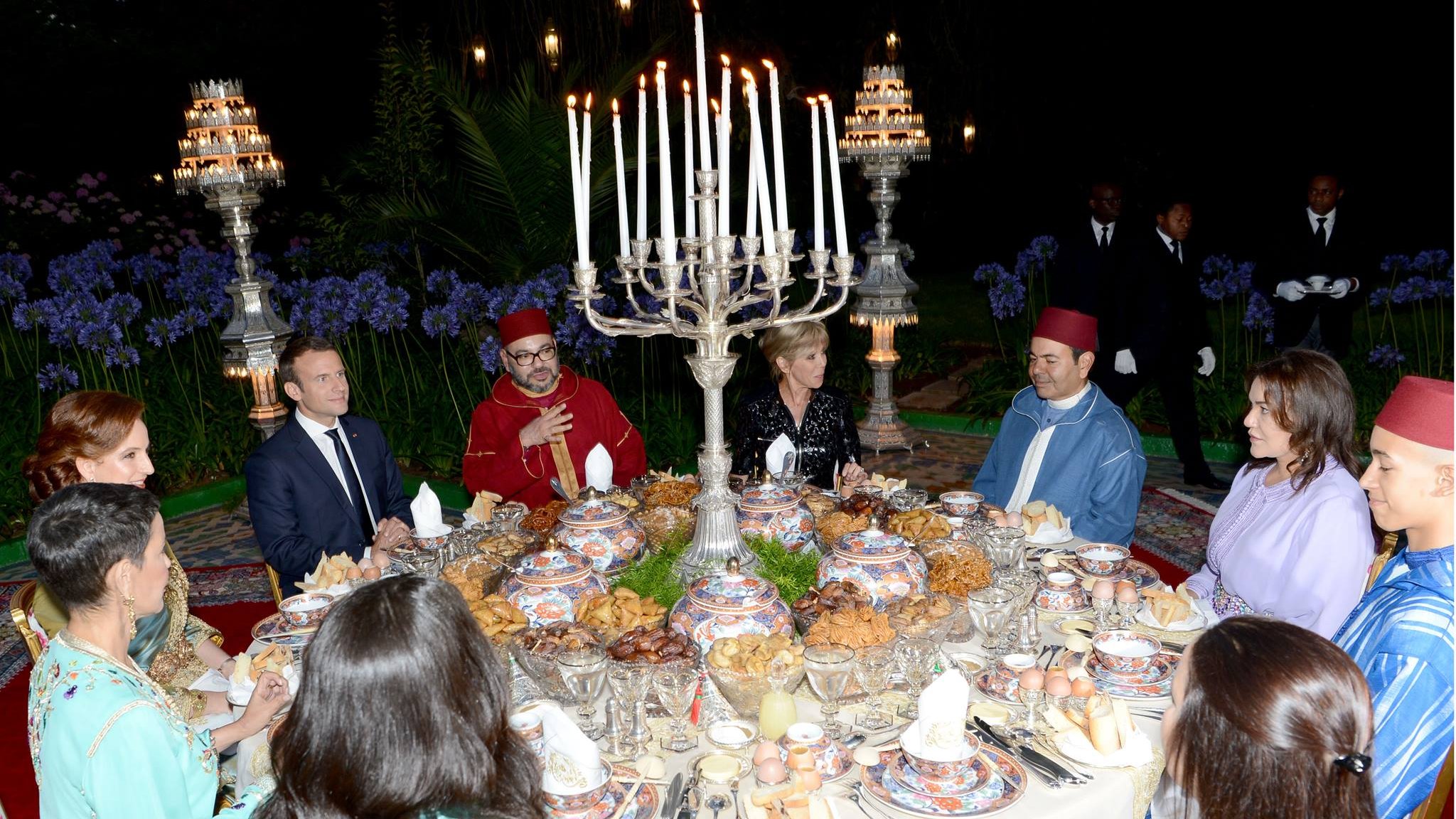 تظهر الصورة مجموعة من الأشخاص حول مائدة رائعة مُعدّة لوجبة طعام فاخرة. المائدة مزينة بأطباق متعددة تحتوي على أطعمة متنوعة، وتحيط بها شمعدانات كبيرة وأجواء أنيقة. يرتدي بعض الضيوف ملابس تقليدية مغربية، بينما يرتدي آخرون ملابس رسمية. تبدو الصورة وكأنها تُعبر عن احتفال خاص أو مناسبة اجتماعية، مع خلفية من النباتات والزهور التي تضفي جوًا جميلًا.