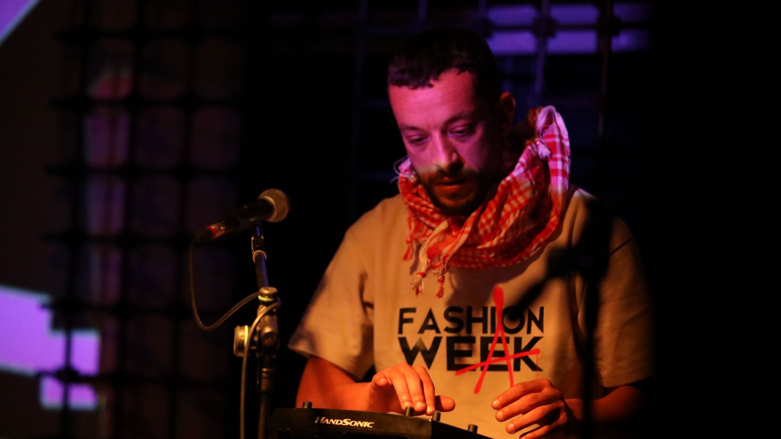 L'immagine mostra un musicista che sta esibendosi su un palco. Indossa una maglietta con la scritta "FASHION WEEK" e una sciarpa a quadri. È concentrato mentre suona un dispositivo elettronico, probabilmente un sintetizzatore o una drum machine. Il palco è illuminato da luci colorate che creano un'atmosfera vivace e coinvolgente. Dietro di lui, si intravedono proiezioni visive che aggiungono dinamismo alla performance.