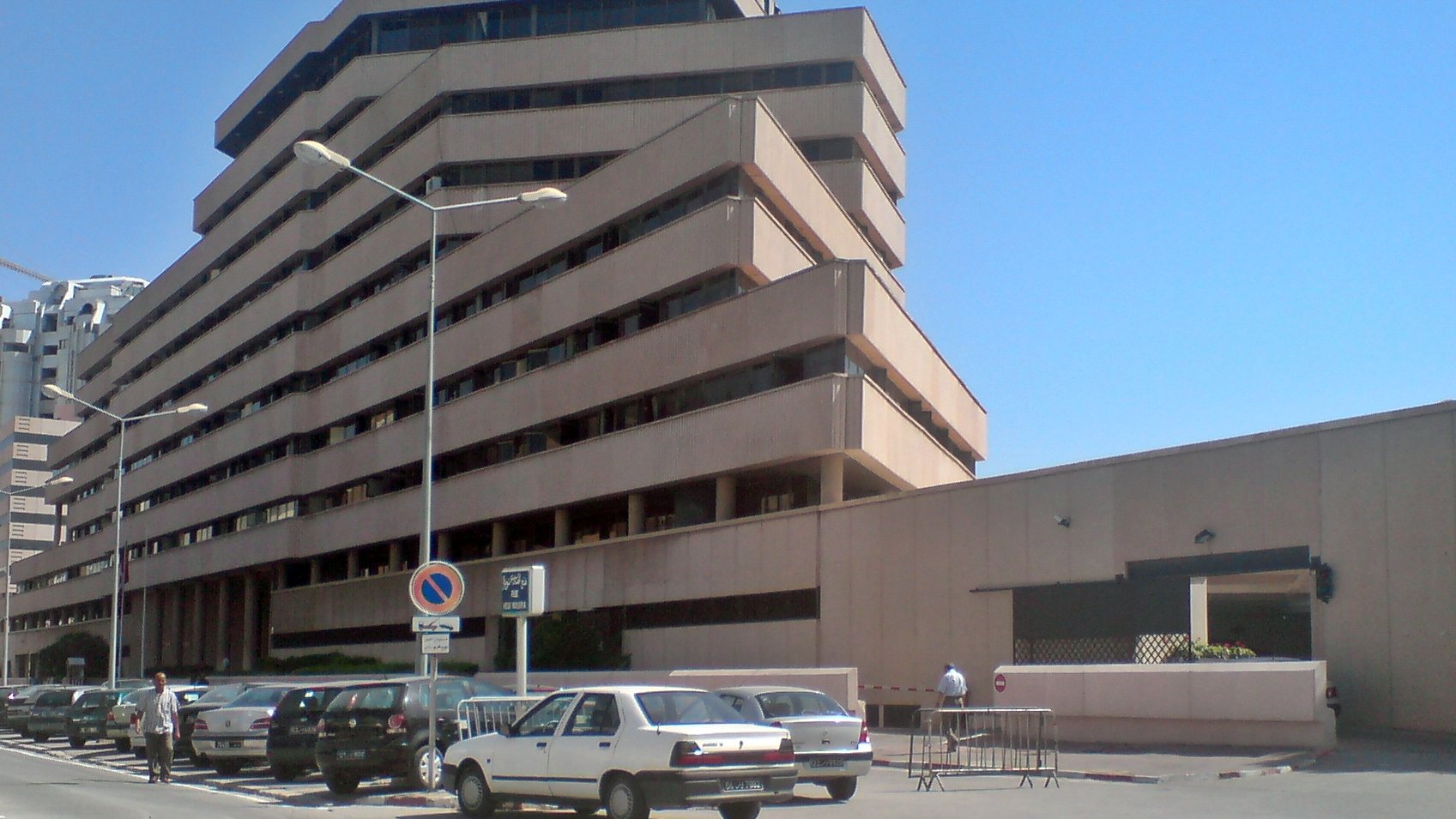 L'image montre un bâtiment moderne à plusieurs étages avec une architecture angulaire et des balcons en saillie. La façade est en béton, avec des couleurs neutres. On peut voir des voitures garées le long de la route, et il semble y avoir des piétons dans la zone. Le ciel est dégagé, ce qui donne une atmosphère ensoleillée à la scène. Le bâtiment dégage une impression de fonctionnalité et de modernité.