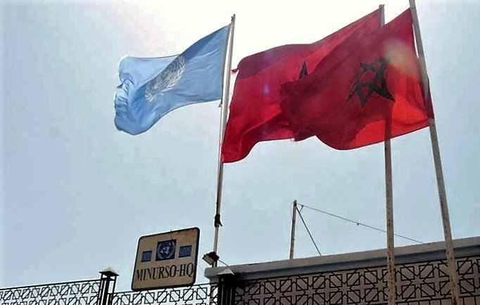 L'image montre deux drapeaux flottant sur des mâts. À gauche, il y a le drapeau de l'ONU, qui est de couleur bleu clair avec un emblème en blanc au centre. À droite, le drapeau du Maroc, qui est rouge avec une étoile verte à cinq branches. En dessous des drapeaux, on peut voir un panneau indiquant "MINURSO HQ", associé à des éléments graphiques de l'Union Européenne. L'ensemble de la scène est éclairé par une lumière vive, suggérant un temps ensoleillé.