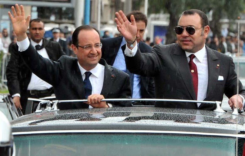 L'image montre deux hommes qui semblent être dans une voiture décapotable. L'un d'eux, à gauche, porte un costume sombre avec une cravate, tandis que l'autre à droite porte un costume avec des accessoires distinctifs. Ils saluent la foule avec la main, semblant sourire et être en bonne humeur. En arrière-plan, il y a des personnes et des voitures, probablement lors d'un événement officiel ou d'une visite. Le temps semble être pluvieux, avec des gouttes d'eau visibles sur la voiture.