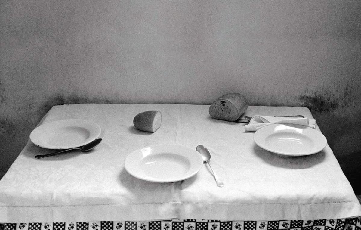 L'image montre une table couverte d'une nappe blanche, où se trouvent deux assiettes blanches, une cuillère et un couteau. Un petit pain est positionné sur un côté et un verre est renversé au centre de la table. L'ambiance générale est sobre et monochrome, ce qui donne une impression de simplicité et de calme. Les murs derrière la table sont légèrement ombragés, ajoutant un effet de profondeur à la composition.