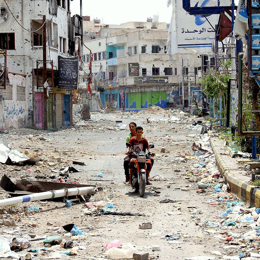 The image depicts a deserted street scene that appears to be in disrepair and heavily littered. There is debris scattered across the ground, suggesting neglect or the aftermath of a conflict. A couple of children can be seen riding a motorcycle down the street, which seems to be a common mode of transportation in the area. Buildings along the street show signs of damage, with faded signs and graffiti on the walls. Overall, the atmosphere conveys a sense of abandonment and deterioration.