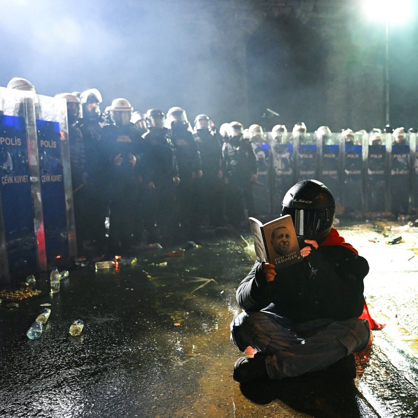 Un manifestant, masqué, lit un livre, face à des policiers en tenue anti-émeute.