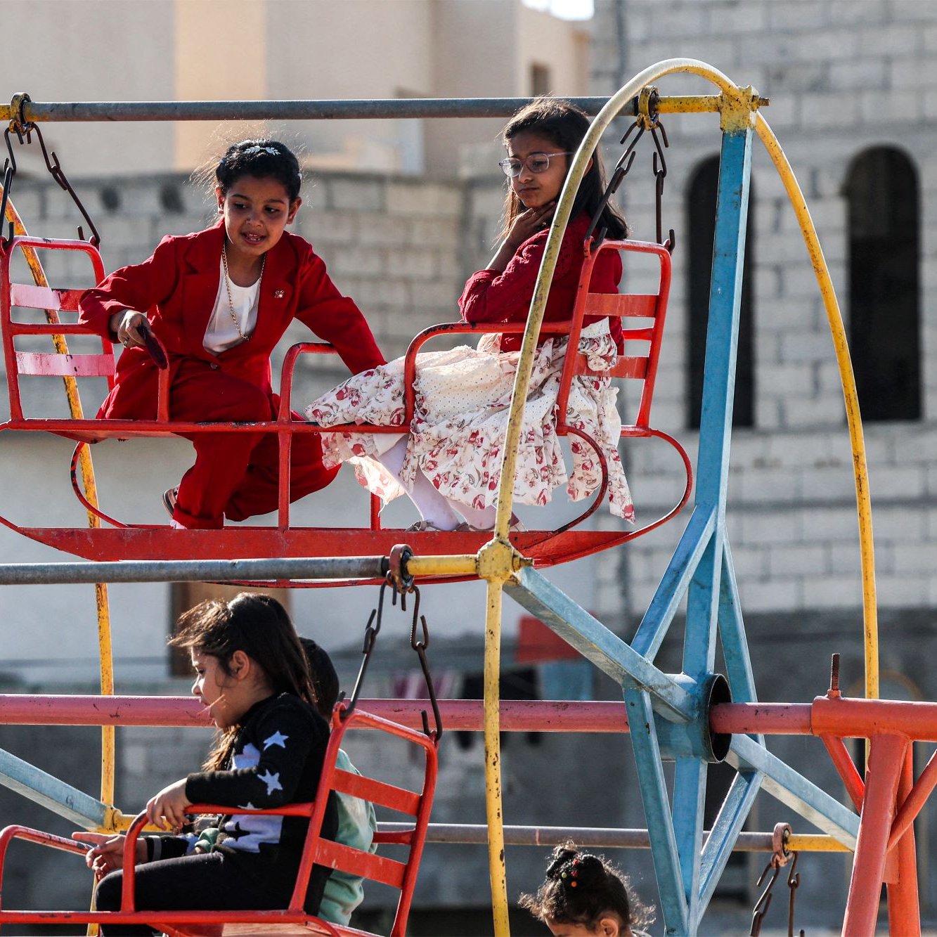 در تصویر، چند کودک در یک وسیله بازی در پارک دیده می‌شوند. وسیله بازی شبیه یک چرخ‌وفلک است و رنگ‌های روشنی دارد. دو کودک در بالای چرخ‌وفلک نشسته‌اند؛ یک پسر با لباس قرمز و یک دختر با لباس سفید و گل‌دار. در پایین نیز تعدادی کودک دیگر در حال بازی و نشستن هستند. فضای تصویر دارای حس شادی و نشاط است و نشان‌دهنده لحظات خوشی از دوران کودکی است. زمینه تصویر ساختمان‌هایی با نمای ساده به چشم می‌خورد.