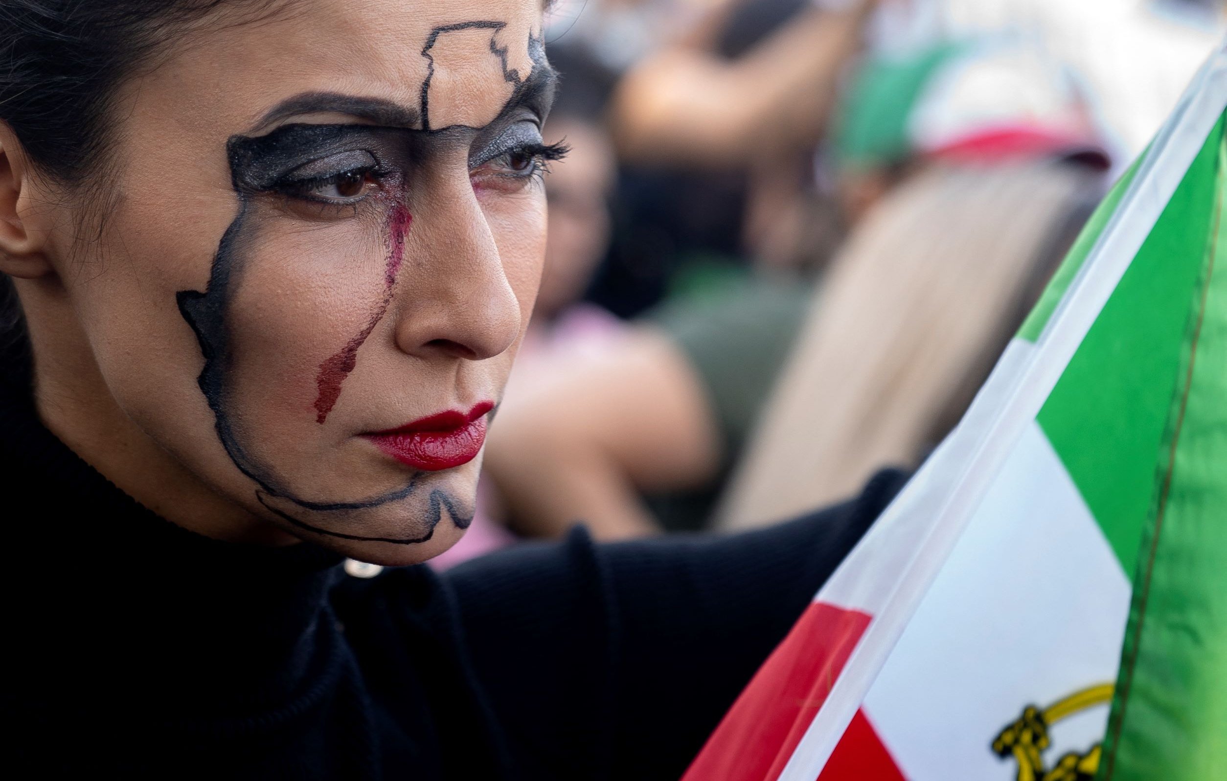 L'immagine mostra una donna con un trucco facciale elaborato, che rappresenta una mappa del suo paese. Ha un'espressione seria e intensa, con un tratto rosso che scende lungo il suo viso, suggerendo emozione o una narrazione profonda. In una mano tiene una bandiera, evidenziando un forte senso di identità nazionale e di protesta. Sullo sfondo si intravedono altre persone, creando un'atmosfera di raduno o manifestazione.
