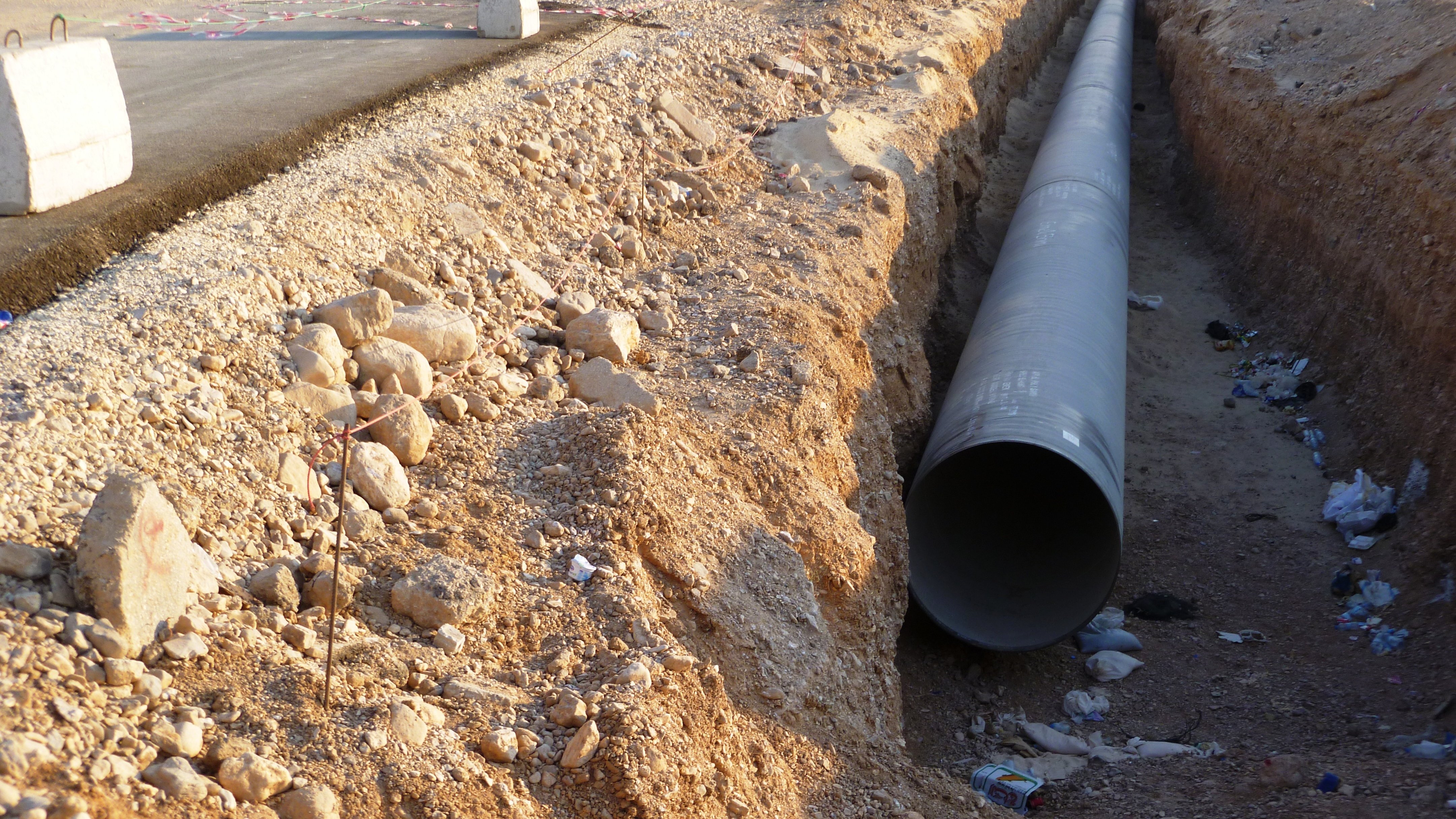 تظهر الصورة مشهدًا لموقع عمل حيث تم حفر خندق في أرض جافة. في وسط الخندق، يوجد أنبوب كبير على الأرض، ويبدو أنه يتم إعداده للتركيب. يحيط بالمنطقة بعض الحصى والصخور، مما يدل على أعمال البناء. في الخلفية، يمكن رؤية معدات بناء تظهر أنها تعمل في الموقع. كما يوجد بعض المخلفات والقمامة حول الخندق، مما يشير إلى تواجد نشاط بشري.