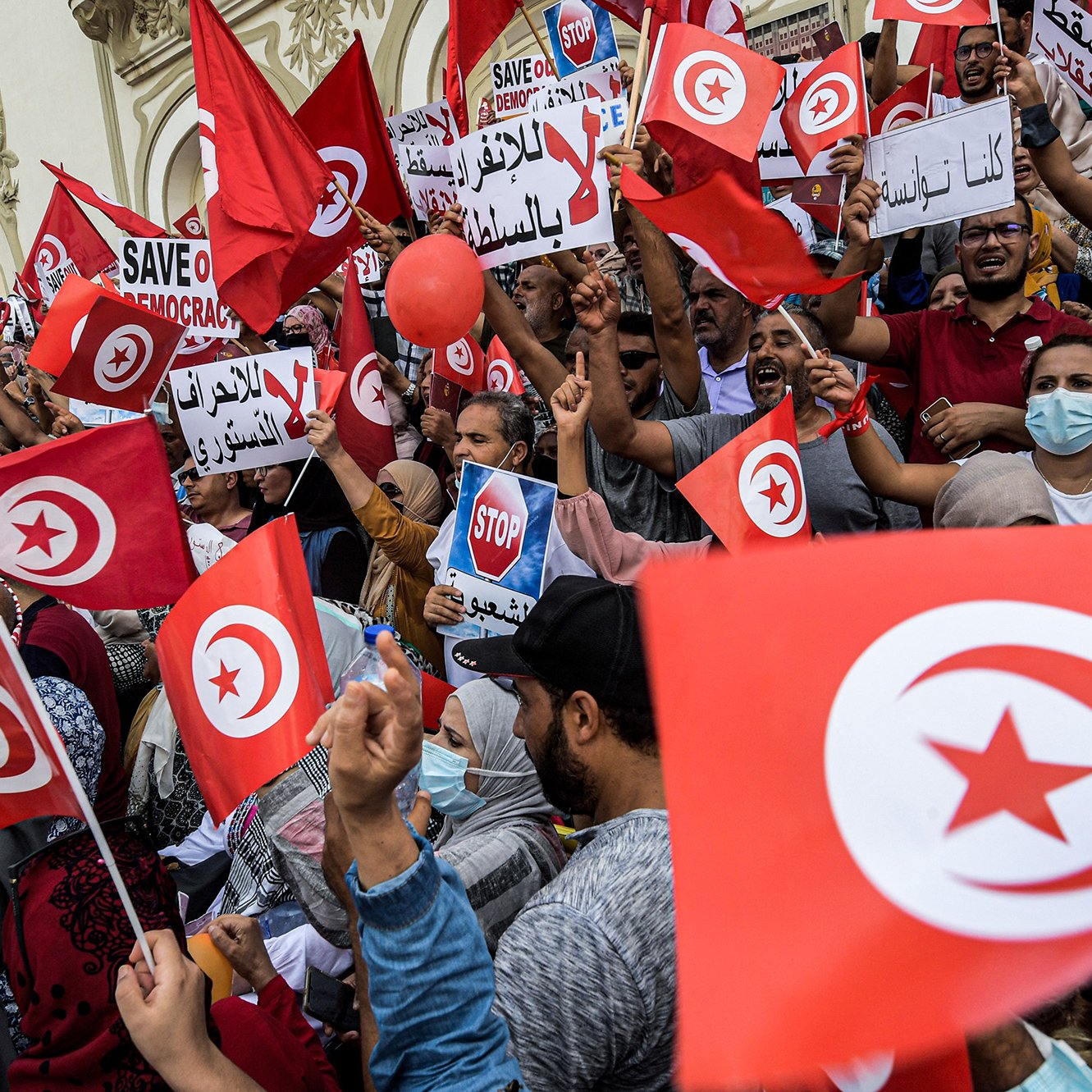 L'image montre une foule manifestante en Tunisie, brandissant des drapeaux tunisiens et des pancartes. Les participants expriment des messages en faveur de la démocratie, avec des slogans tels que "Sauvez la démocratie" et "Stop". On peut voir des gens de différents âges et genres, certains portant des masques. L'ambiance semble énergique et engagée, avec un fort esprit de revendication collective.