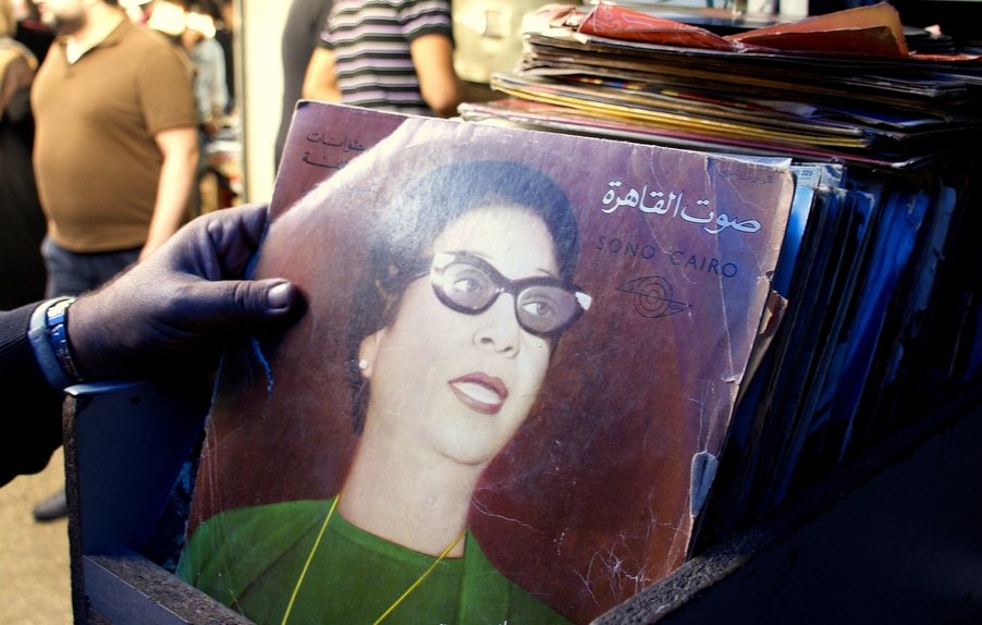 L'image montre une personne tenant un disque vinyle dans un marché. Le disque est de couleur foncée, avec une illustration d'une femme portant des lunettes et des vêtements vintage. En arrière-plan, on aperçoit d'autres personnes dans un environnement animé, probablement un marché ou une braderie, avec divers objets et éléments visibles autour. L'ambiance semble nostalgique, soulignant l'importance de la musique et de la culture visuelle.