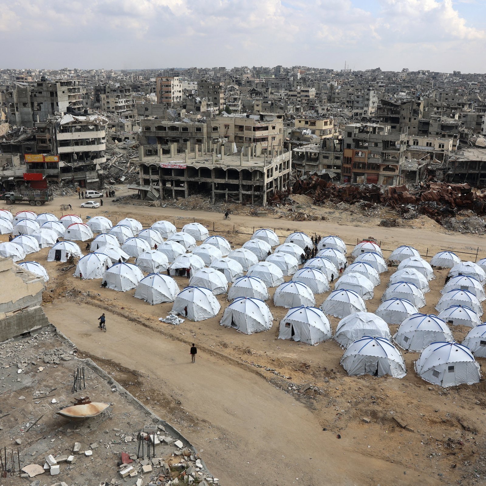 L'image montre une vaste zone de tentes blanches installées sur un terrain aride, entourée de bâtiments en ruines. Les tentes semblent former un camp, probablement pour des personnes déplacées ou des réfugiés. En arrière-plan, on peut voir des structures détruites, ce qui indique un environnement de conflit ou de catastrophe. Le ciel est nuageux, et l'atmosphère générale évoque la désolation et la nécessité d'abri.