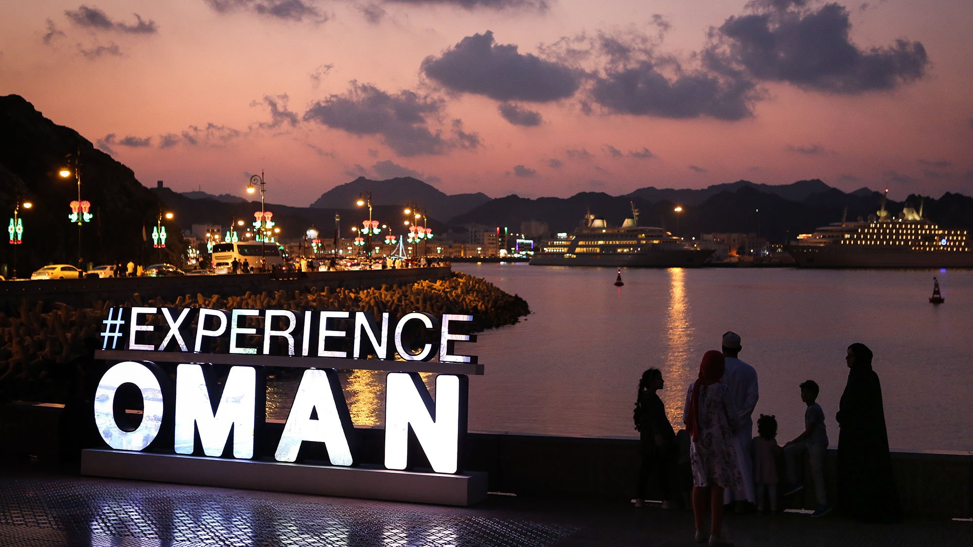 L'image montre une scène au coucher du soleil à Oman, avec des montagnes en arrière-plan et un port où se trouvent des bateaux et des navires de croisière. Au premier plan, il y a un grand panneau lumineux affichant le mot "EXPERIENCE" suivi d'"OMAN". Des personnes se tiennent devant le panneau, admirant la vue sur l'eau. La lumière douce du crépuscule et l'ambiance paisible de la scène créent une atmosphère accueillante.