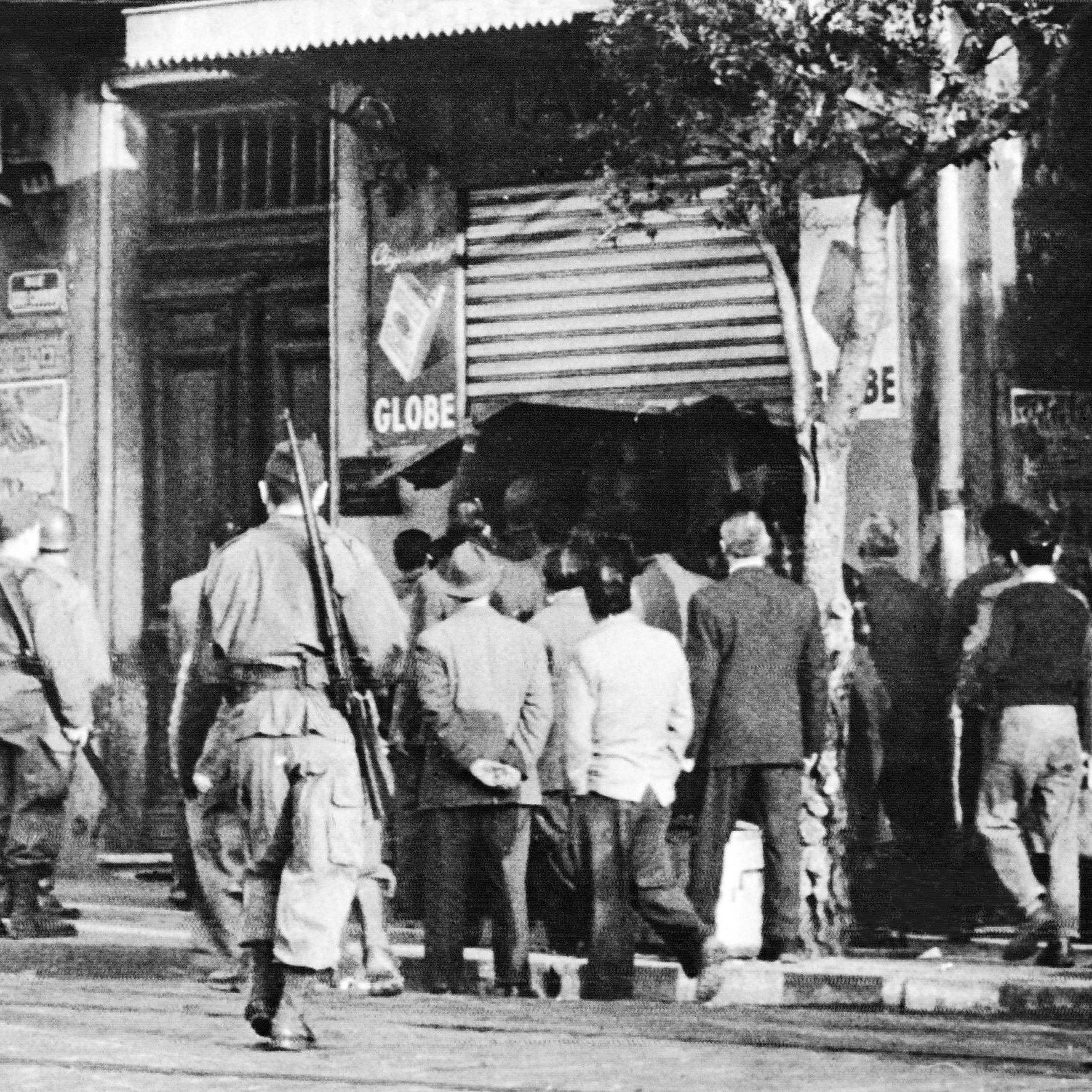 در این تصویر، گروهی از مردم در خیابان در حال نظارت به یک مکان هستند. تعدادی سرباز در نزدیکی آن‌ها ایستاده‌اند که به نظر می‌رسد در حال کنترل اوضاع هستند. این تصویر احتمالاً از یک زمان تاریخی گرفته شده است و نشان‌دهنده تنش یا رویدادی خاص در آن زمان است. حالت افراد و لباس‌هایشان به این احساس دامن می‌زند. برخی از افراد به سمت مغازه‌ای در حال حرکت هستند، در حالی که دیگران از دور نظاره‌گر هستند.