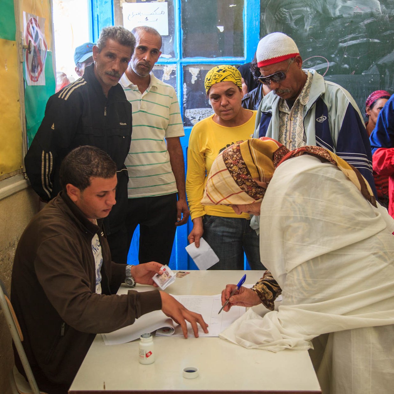 L'image montre un groupe de personnes se rassemblant autour d'un bureau dans une salle. Un homme est assis à une table, où il semble aider une femme, qui est en train de signer un document. Elle porte une tenue claire et un foulard. D'autres personnes, dont des hommes et des femmes, observent la scène, manifestant un intérêt. Les murs sont décorés de couleurs vives, et une fenêtre ouverte laisse entrer la lumière. L'environnement semble informer d'une activité communautaire, peut-être liée à un vote ou à une inscription.